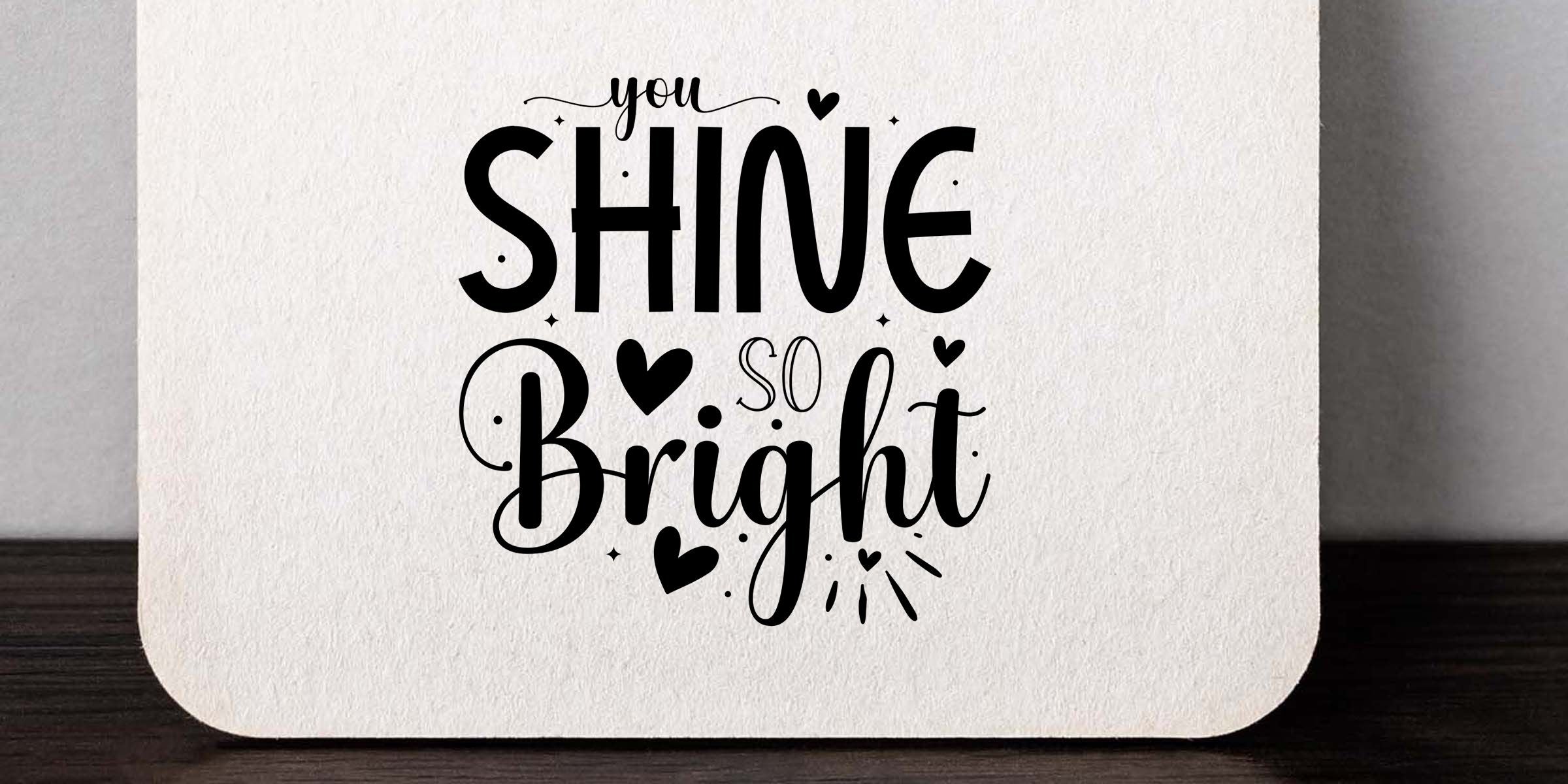 White ceramic coaster with black inspirational typography reading “you shine so bright,” accented by hearts and decorative flourishes on a textured background.