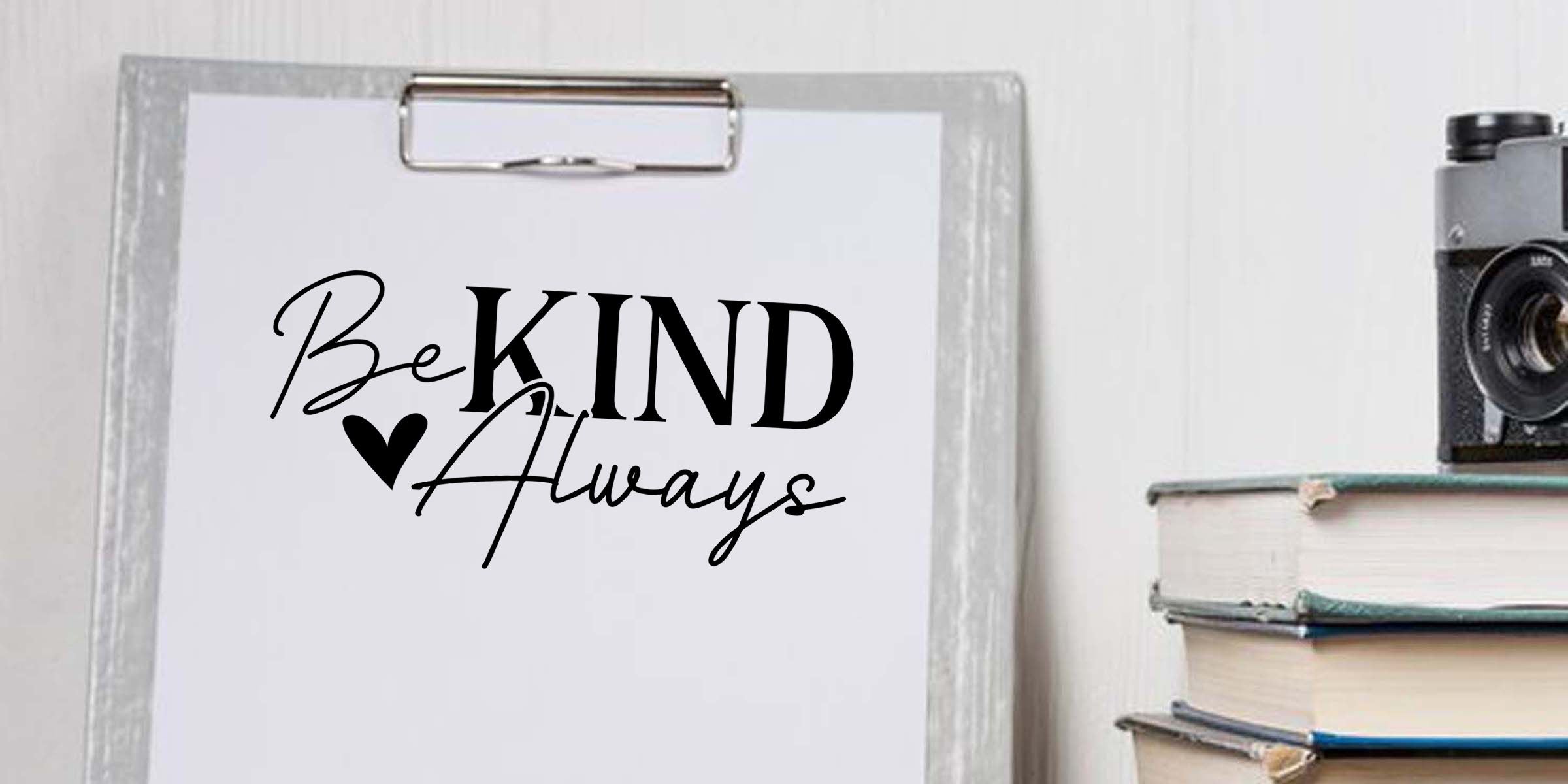Clipboard with “Be Kind Always” typography print, stacked books, and vintage camera in a minimalist decor setting