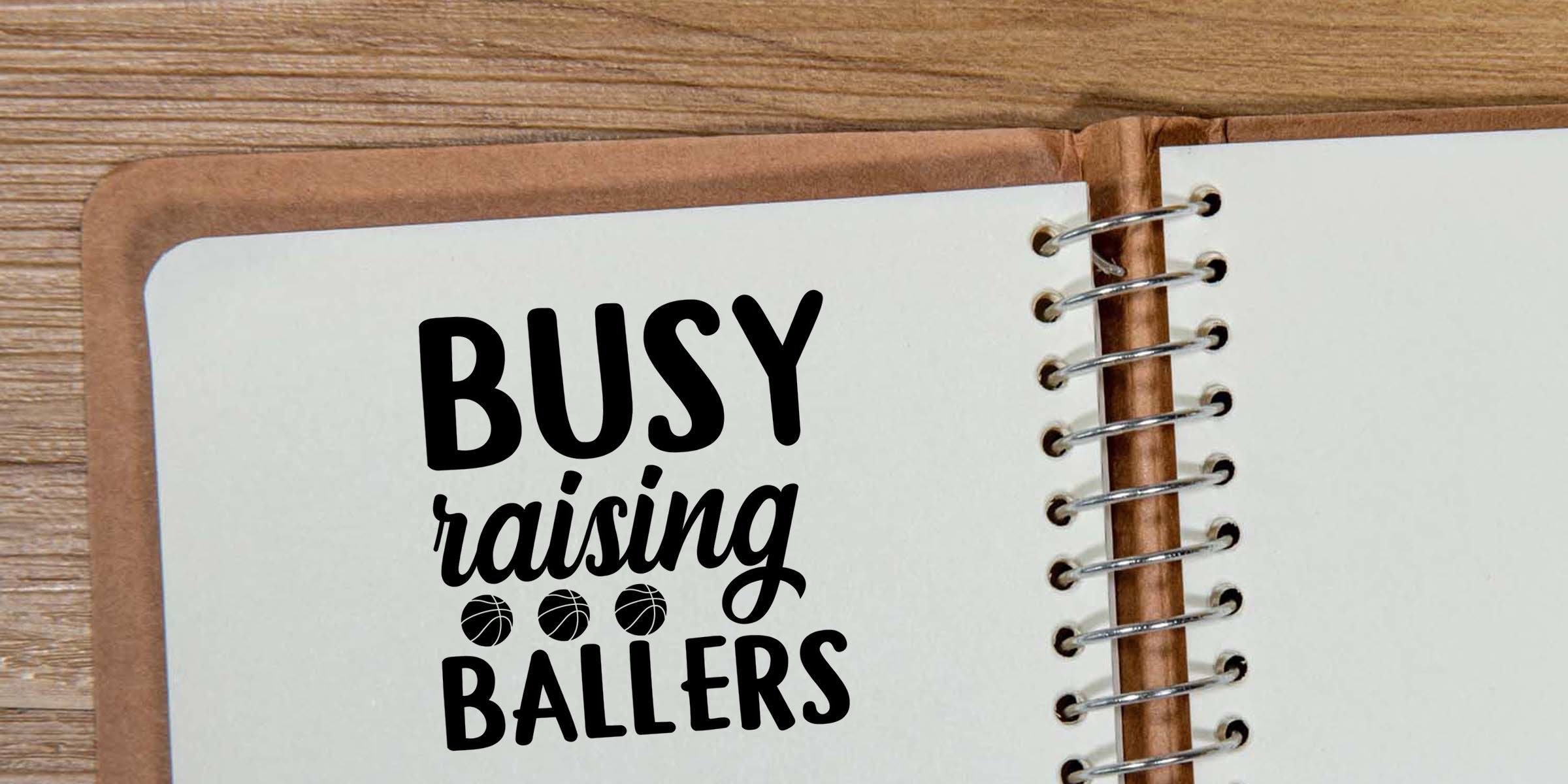 Open spiral notebook on wooden table with “Busy Raising Ballers” text and basketball icons, family basketball mom gift design