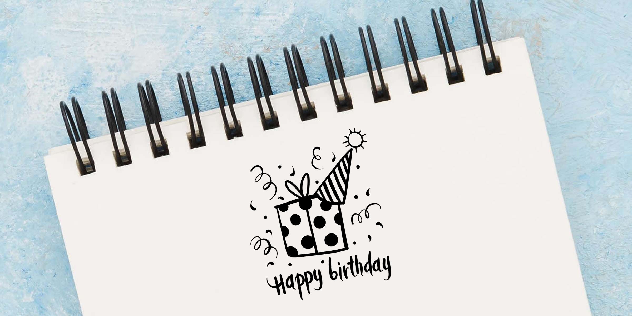 Spiral notebook with happy birthday gift illustration on a white cover against a blue textured background