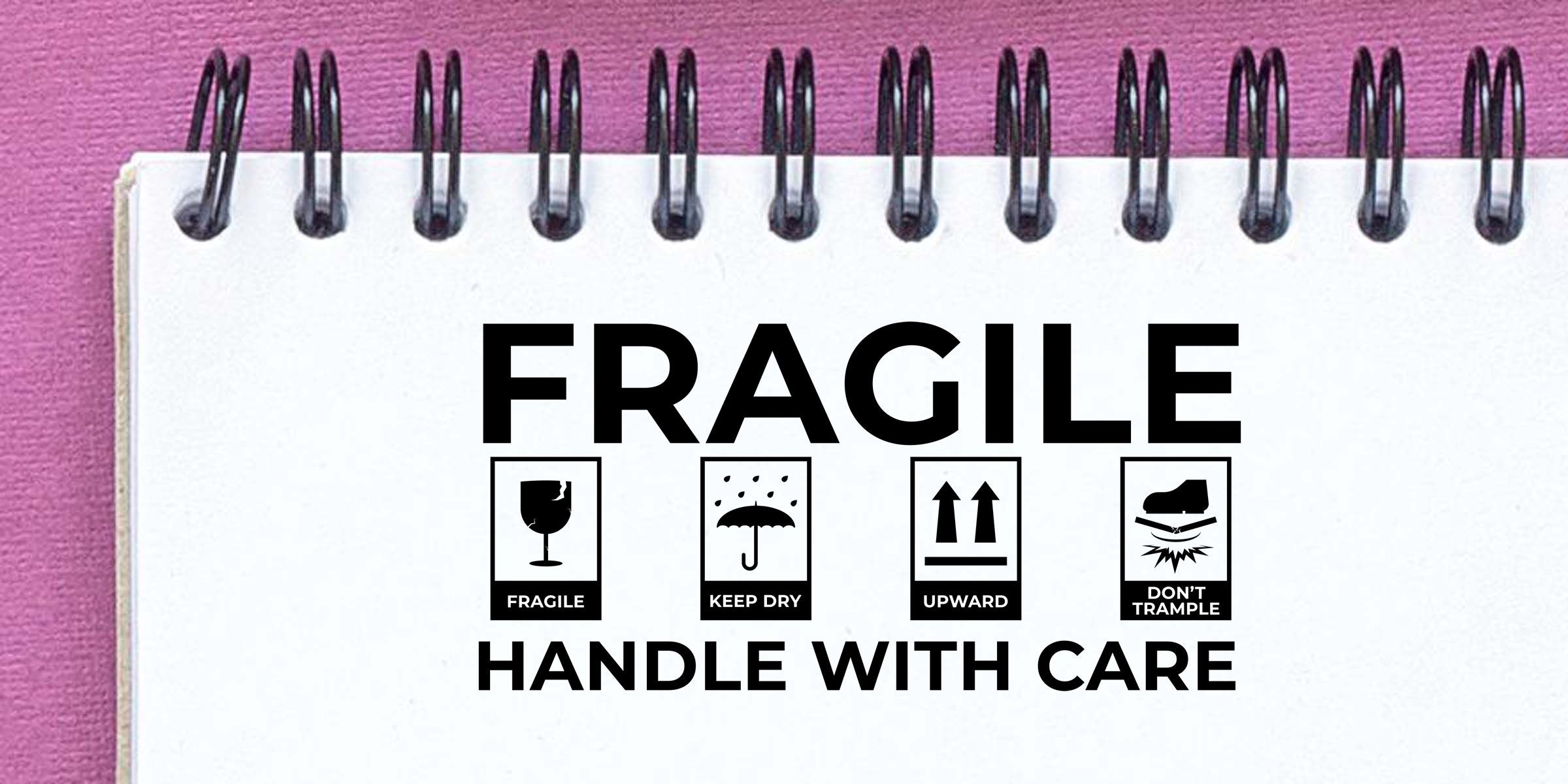 Fragile handle with care packaging warning printed on a spiral notebook page, featuring keep dry, upward, and don’t trample symbols on a pink background