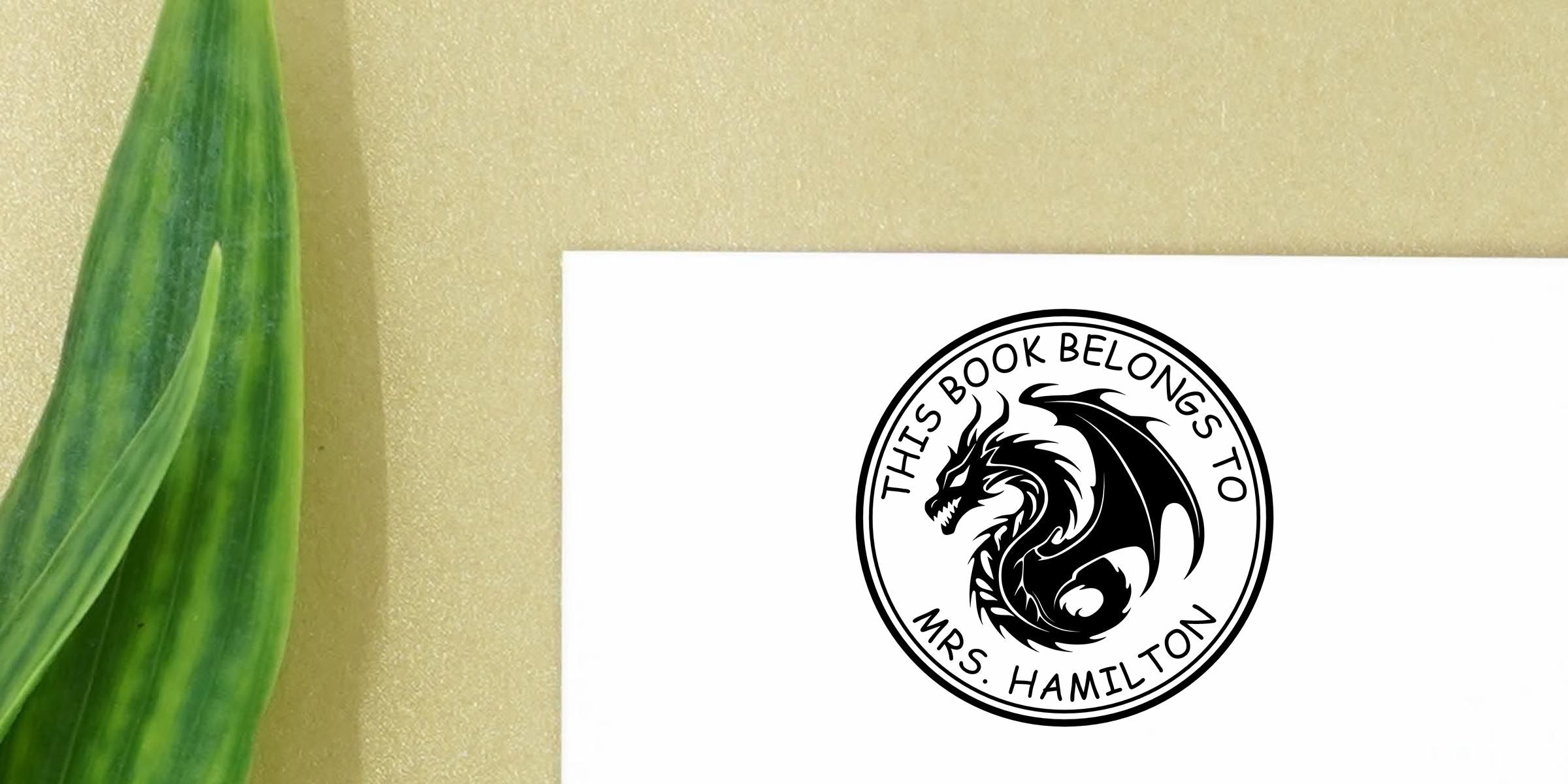Custom dragon book plate label reading “This Book Belongs to Mrs. Hamilton” beside a green decorative leaf on a gold background
