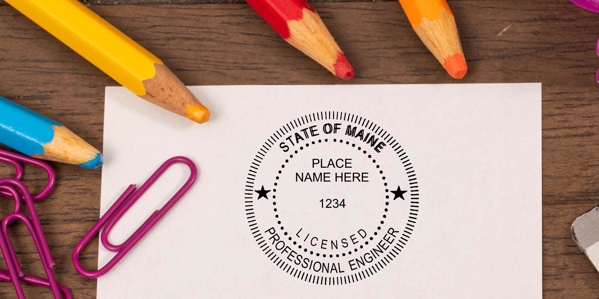 State of Maine licensed professional engineer stamp on white paper surrounded by colored pencils and paper clips on a wooden desk.