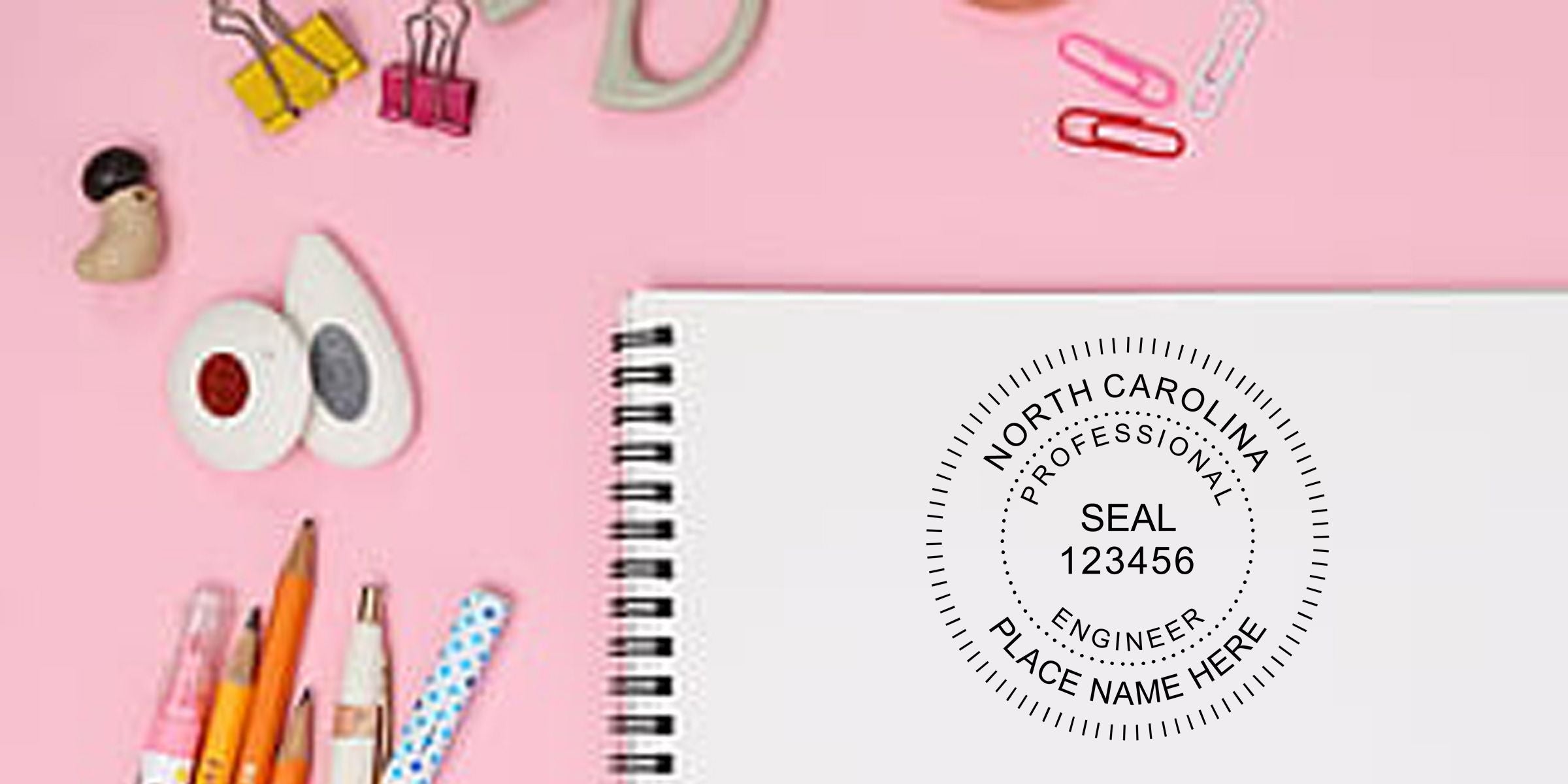 Custom North Carolina professional engineer seal on white spiral notebook surrounded by pens, pencils, binder clips, and stationery on a pink background
