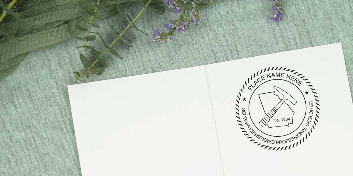 Georgia geologist license stamp with registered professional geologist seal, rock hammer, and state outline on white paper, placed on green fabric with purple flowers, showcasing Georgia geologist stamp requirements.