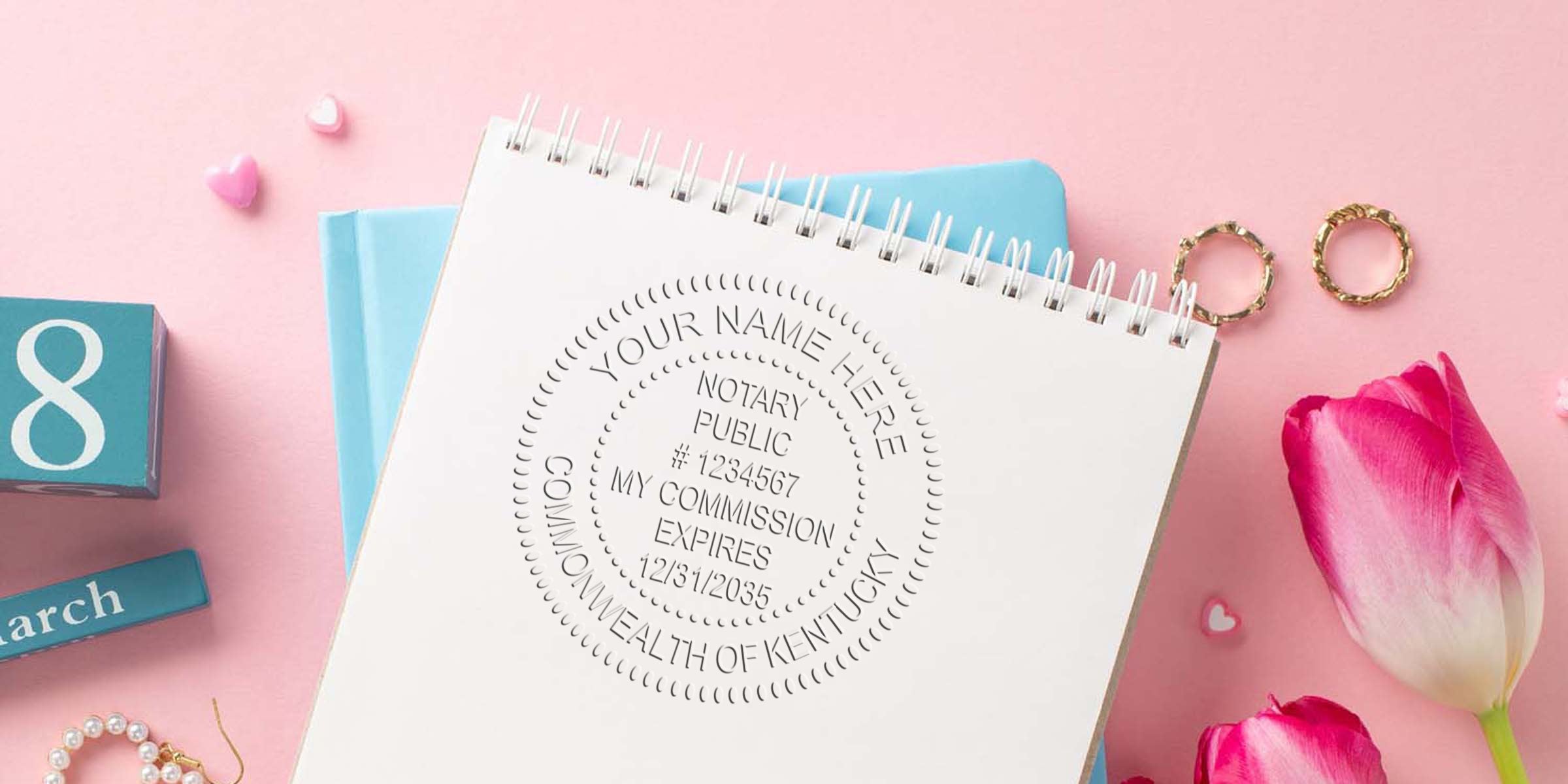 Kentucky notary seal on a commission certificate atop a spiral notebook, surrounded by gold rings and pink tulips on a pastel pink background.