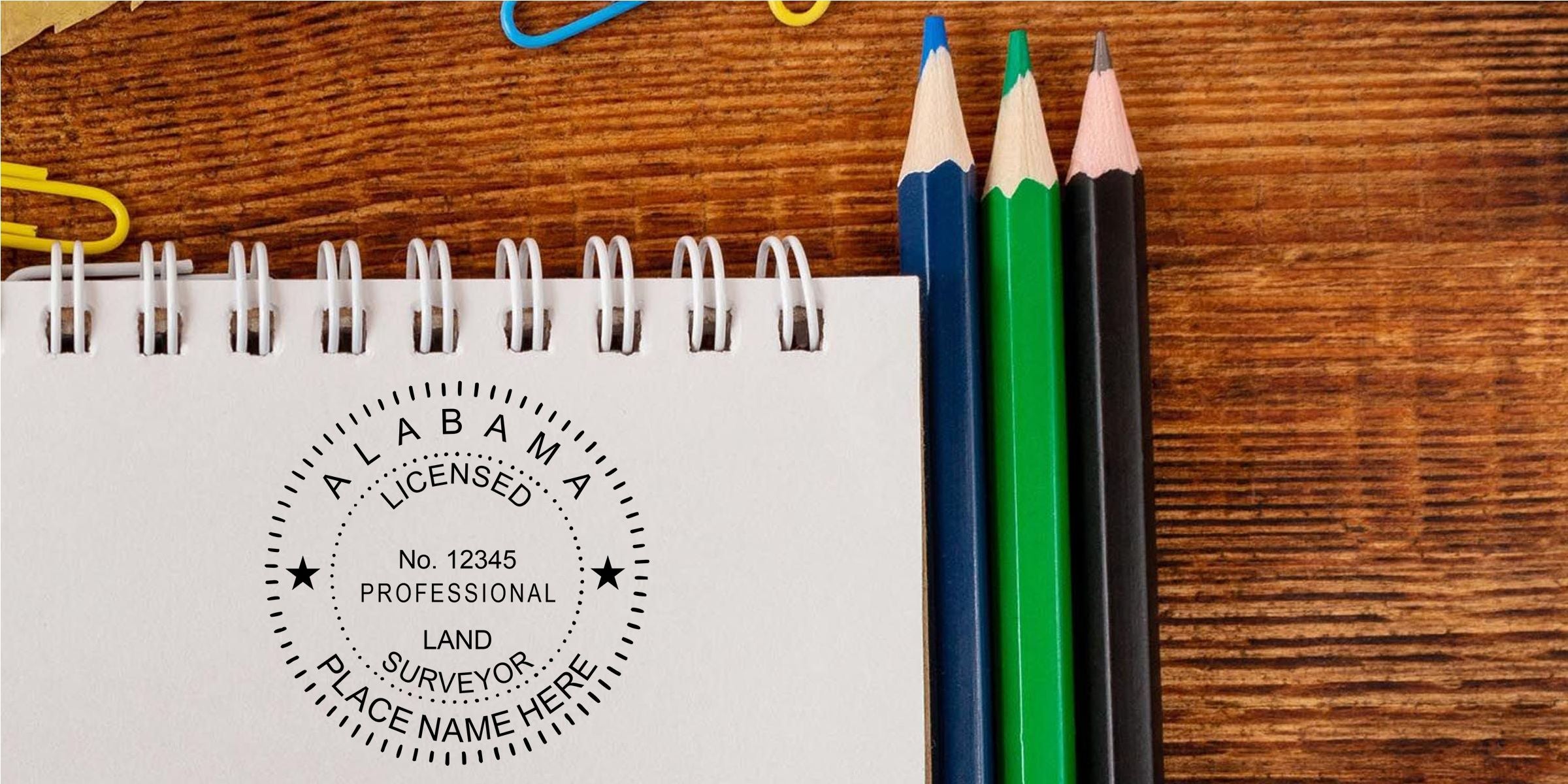 Custom licensed professional land surveyor logo on a spiral notebook beside colored pencils on a wooden desk, with Alabama and place name text for branding and stationery design