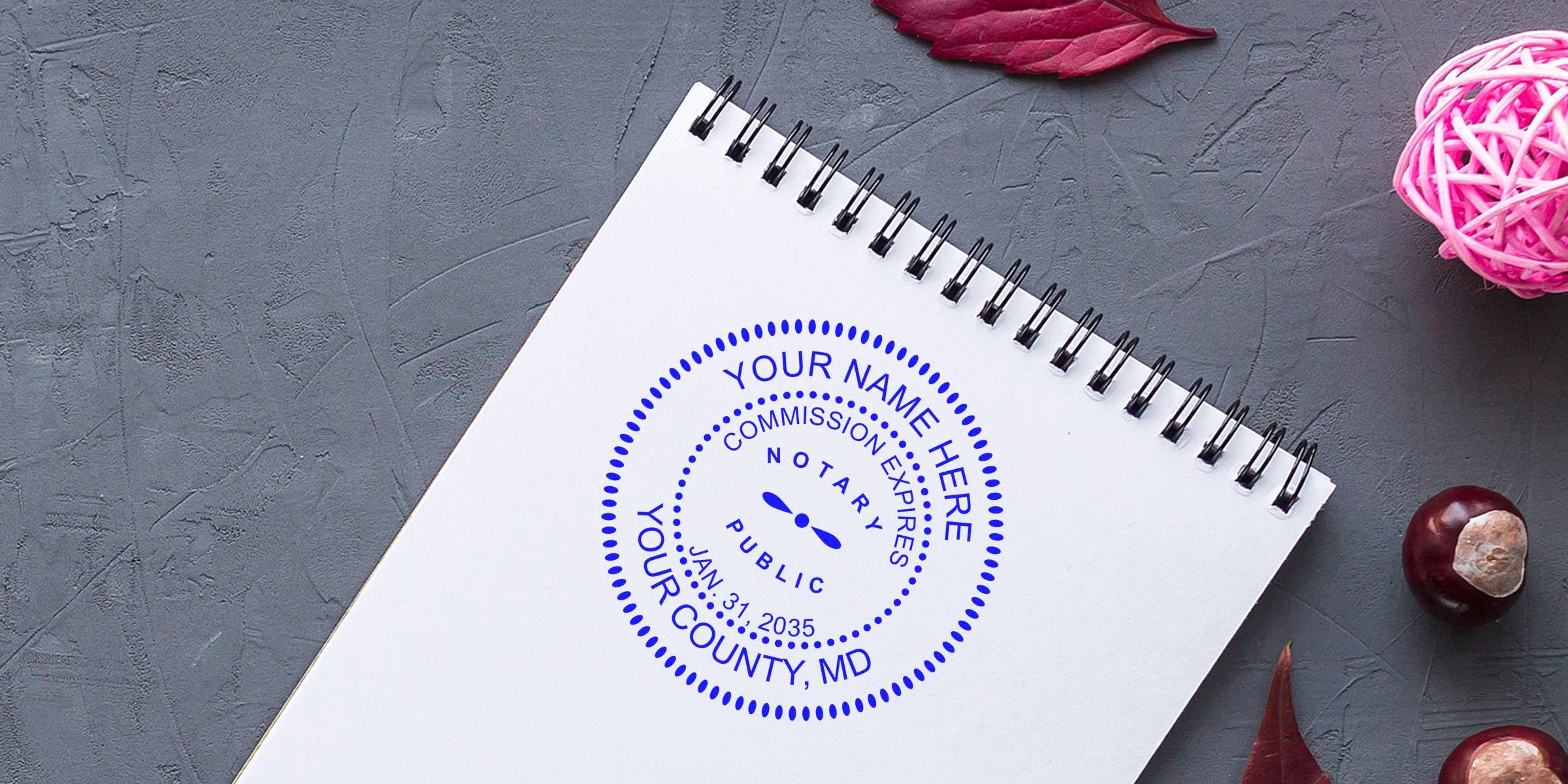 Maryland notary seal custom stamp on white spiral notebook with autumn leaves and chestnuts on gray textured surface, shown in notary stamp guide.
