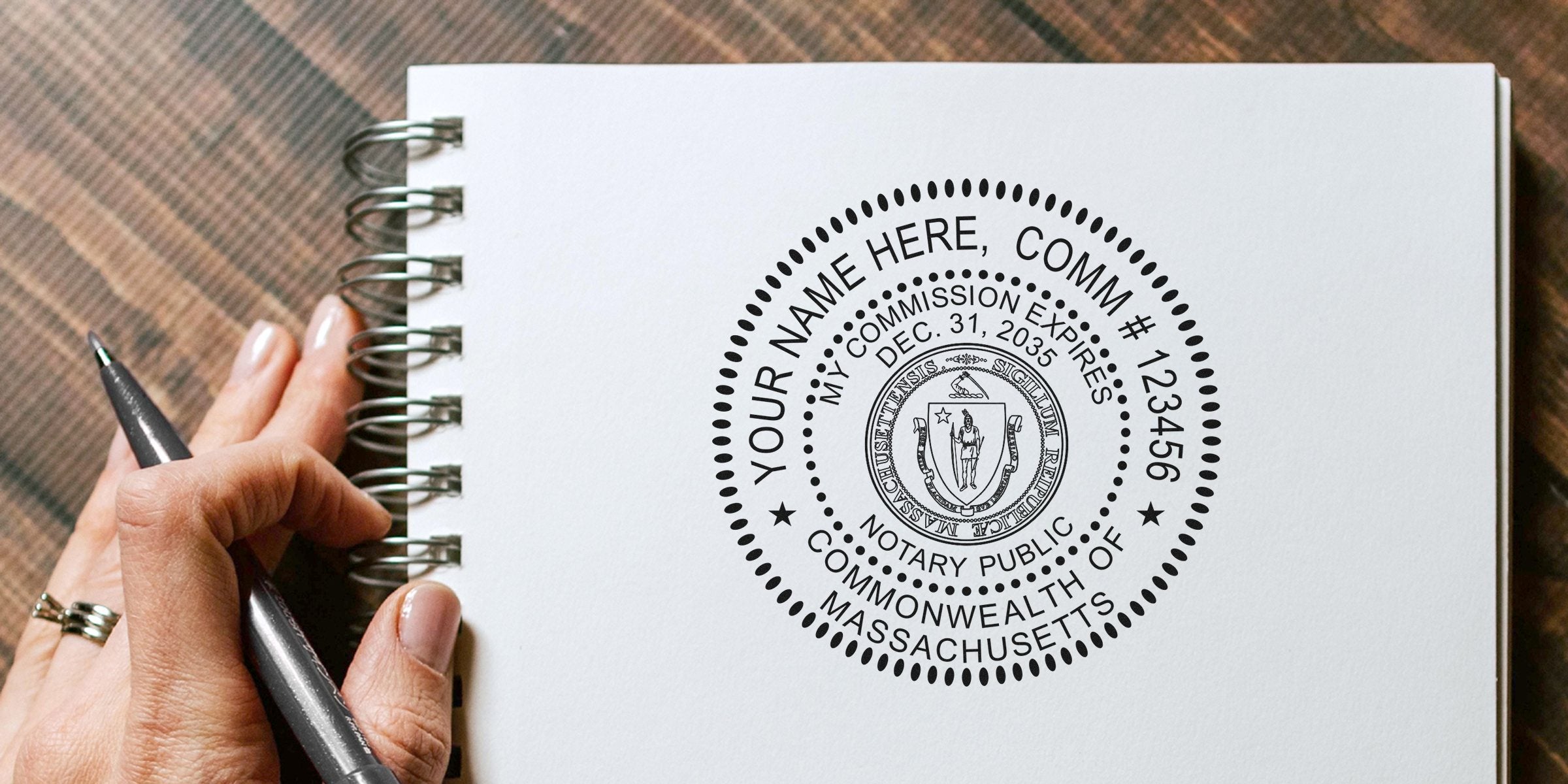 Massachusetts notary public stamp on white spiral notebook with hand holding pen, showing notary commission seal and expiration date, essential notary supplies and tools for Massachusetts.