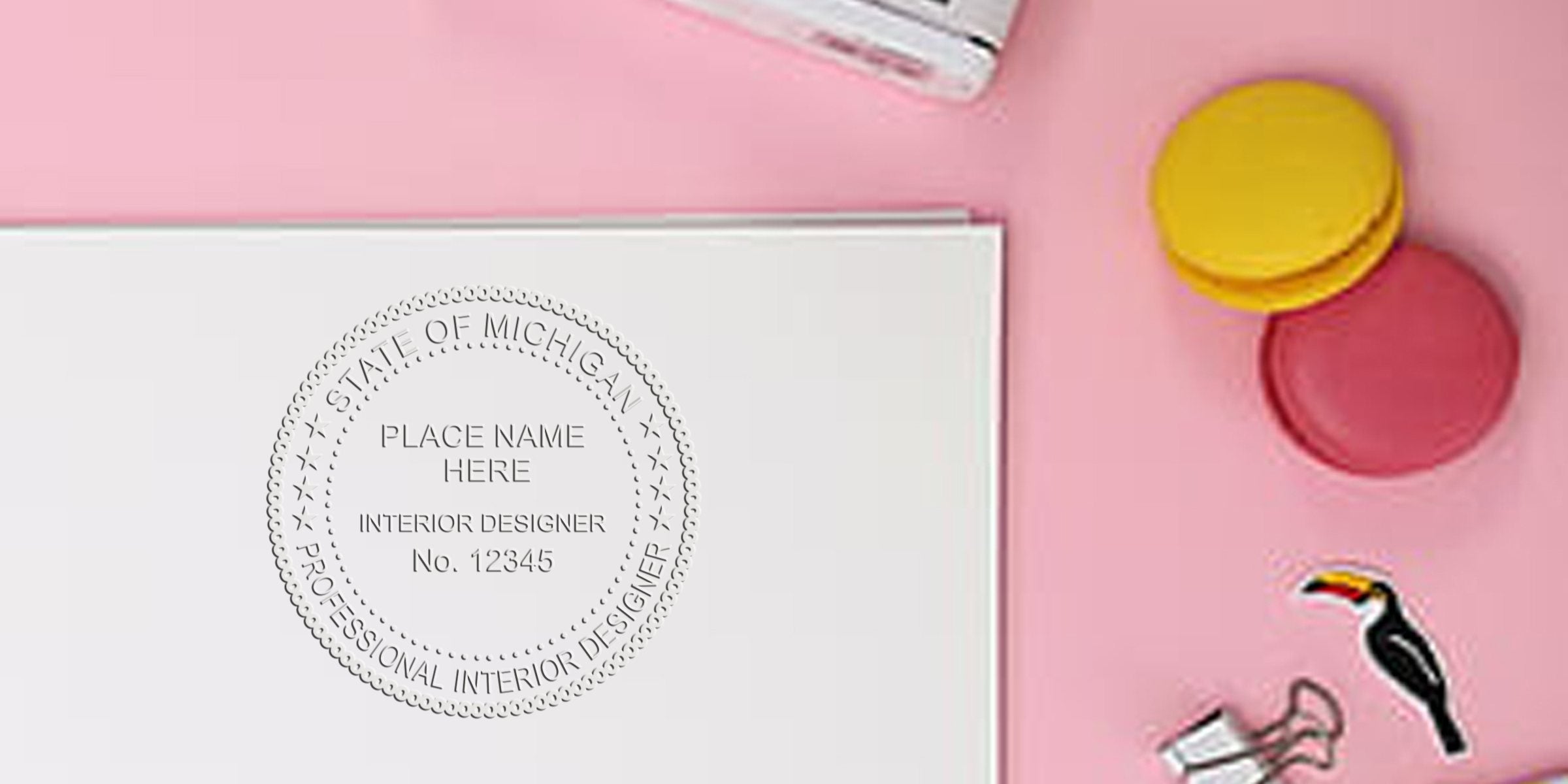 Michigan interior designer’s embossed stamp on white paper, surrounded by colorful macarons and office supplies on a pink background, with quick tips for ordering stamps.