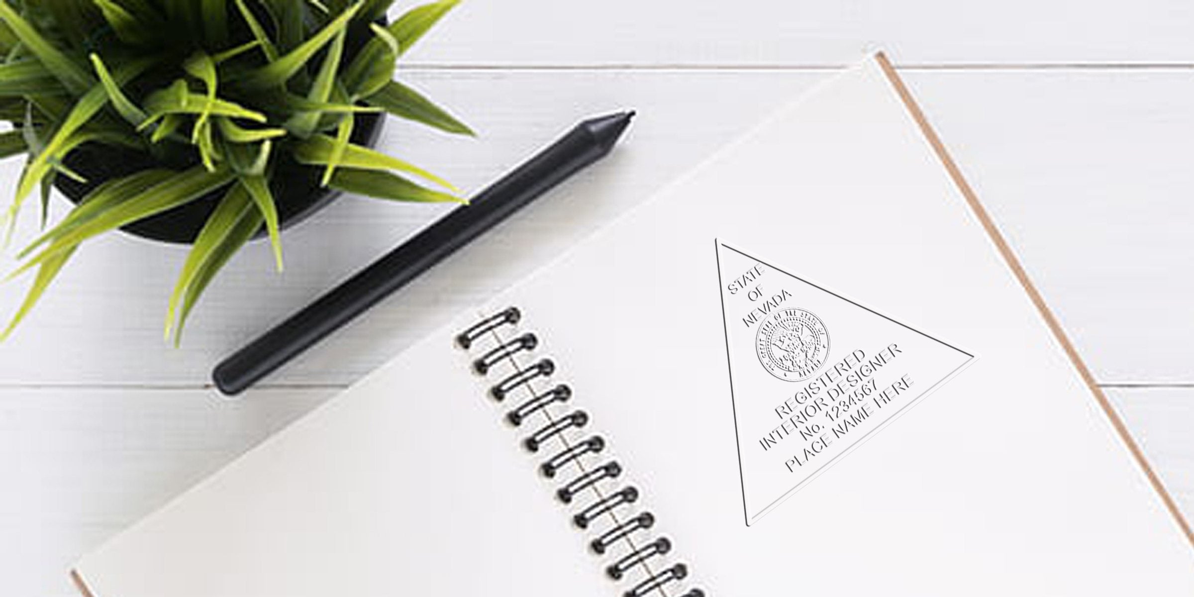 Spiral notebook with Nevada interior designer stamp, black pen, and green plant on white wooden desk for interior design certification and online stamp.