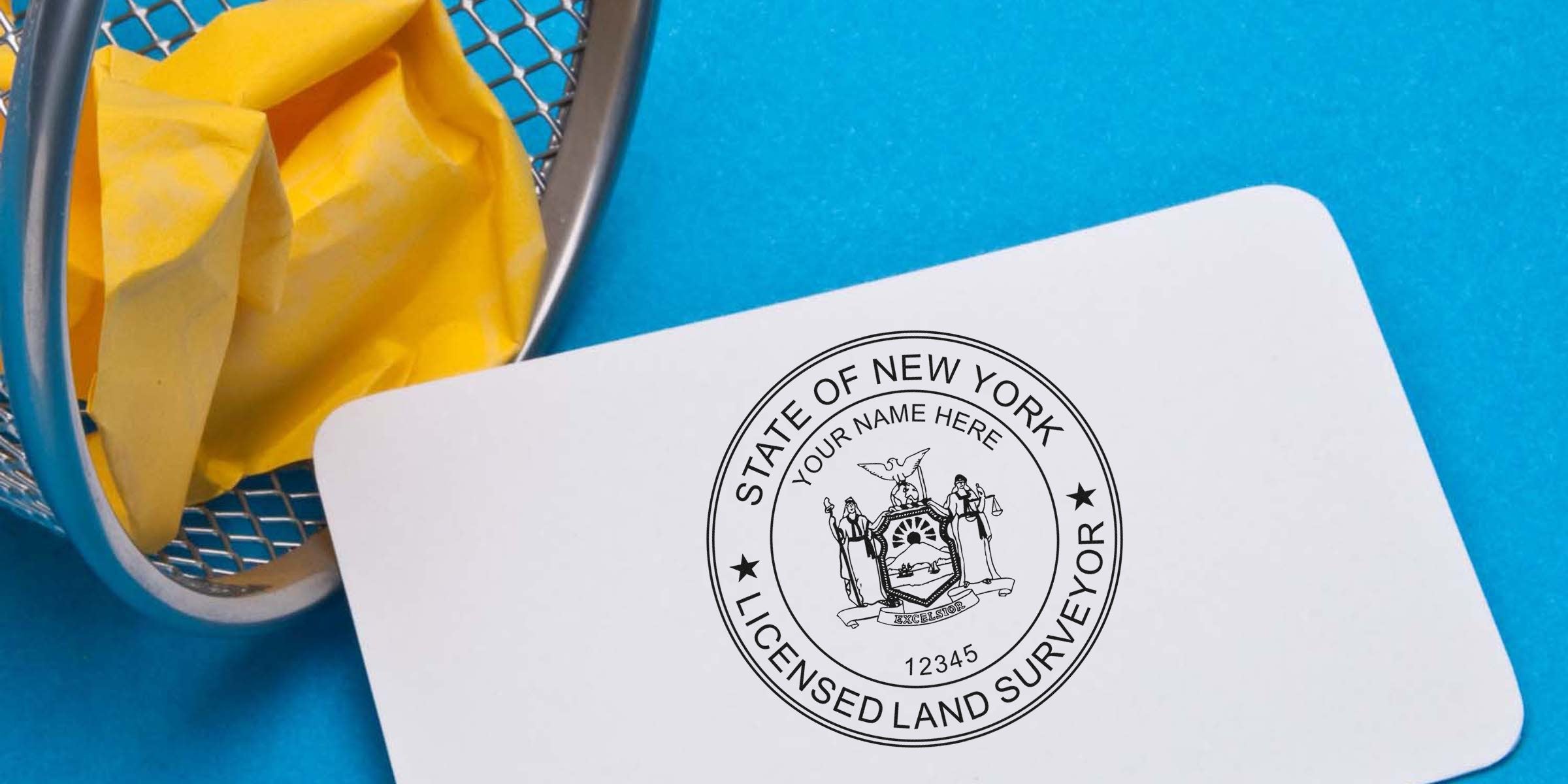 New York land surveyor stamp on white card next to yellow crumpled paper in metal trash bin on blue background, showing land surveyor stamp requirements.