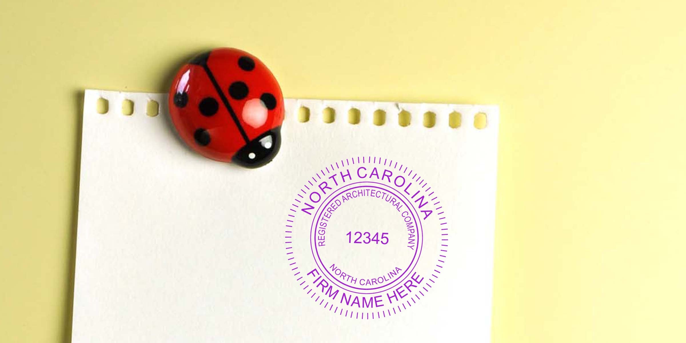 Custom North Carolina architect stamps on white paper, held by a ladybug magnet against a yellow background, showcasing a registered architectural company’s stamp.