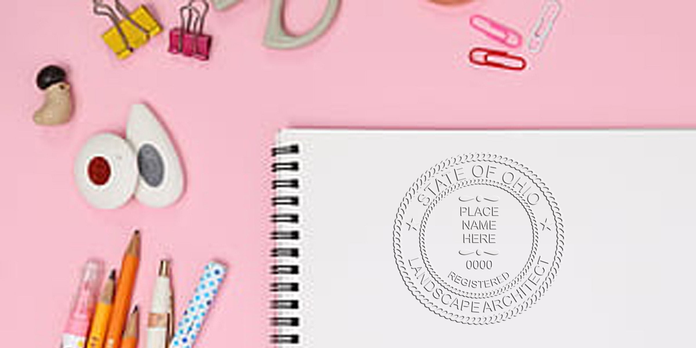 Spiral notebook with Ohio landscape architect seal on pink desk, surrounded by pencils, paper clips, and office supplies, highlighting Ohio landscape architect stamp laws and regulations.