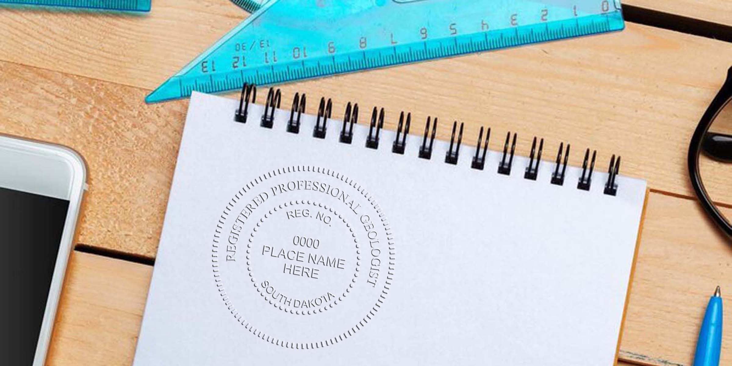 South Dakota geologist seal embossed on white notebook, with blue ruler and pen on wooden desk, using geologist seal embosser.