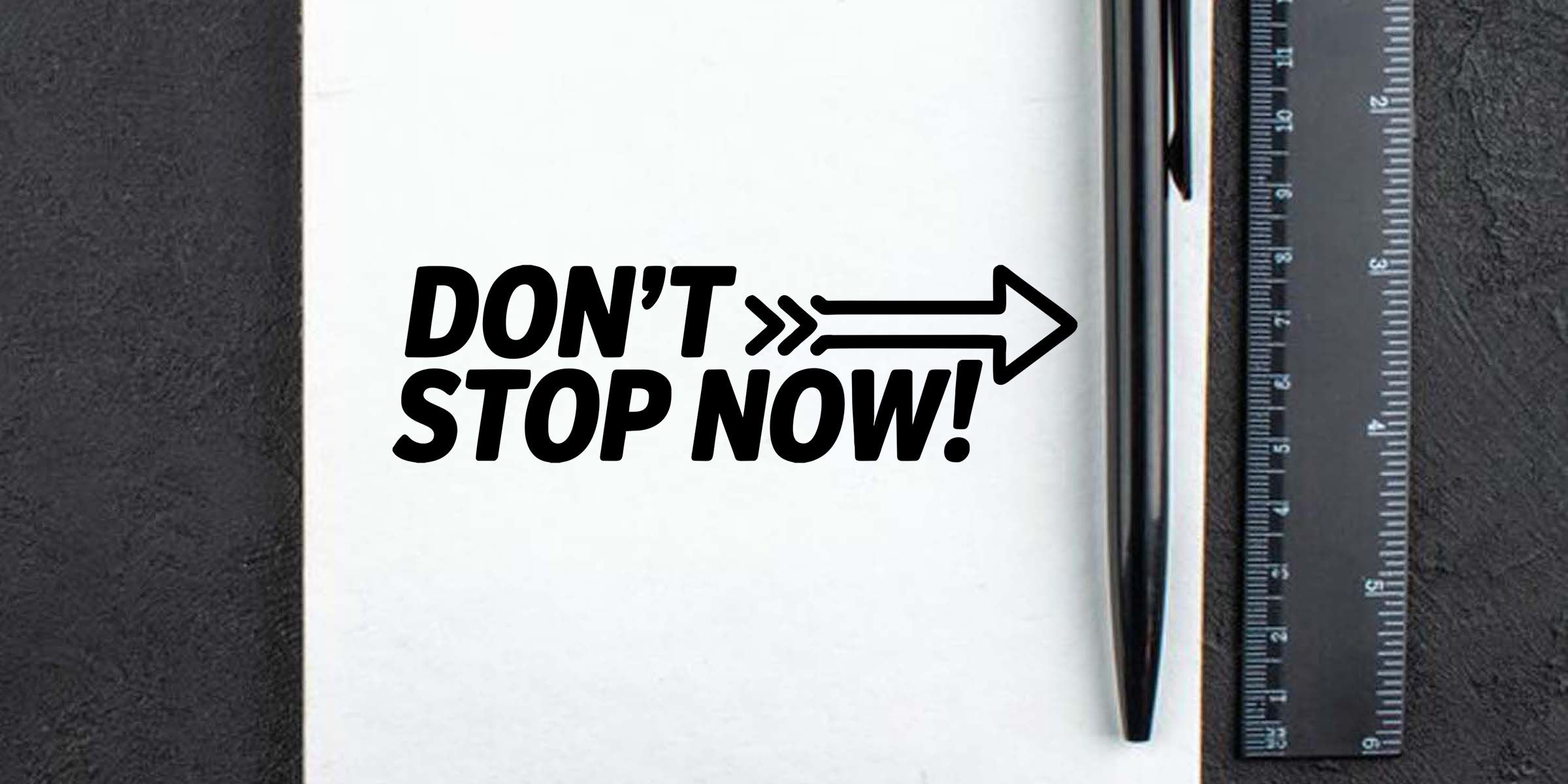 Bold motivational quote "DON'T STOP NOW!" printed on white paper with arrow graphic, beside a pen and ruler on a black textured background