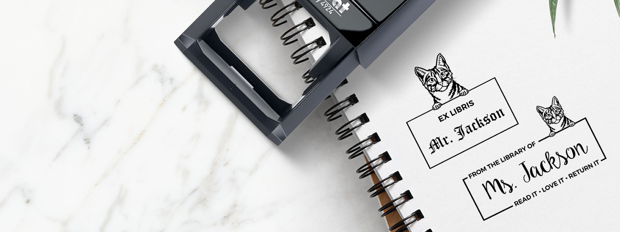 Black stamp tool over a spiral notebook showing Mr. and Mrs. Jackson cat ex libris bookplates on white marble, Egyptian Mau Cat Stamps and Custom Seals.