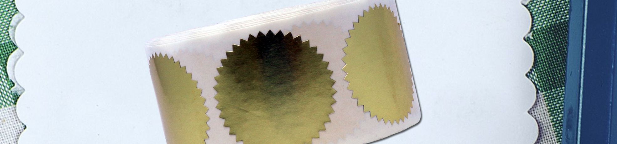 Close-up of gold foil seal stickers with jagged edges on a white scalloped card, featuring Foil Seals.