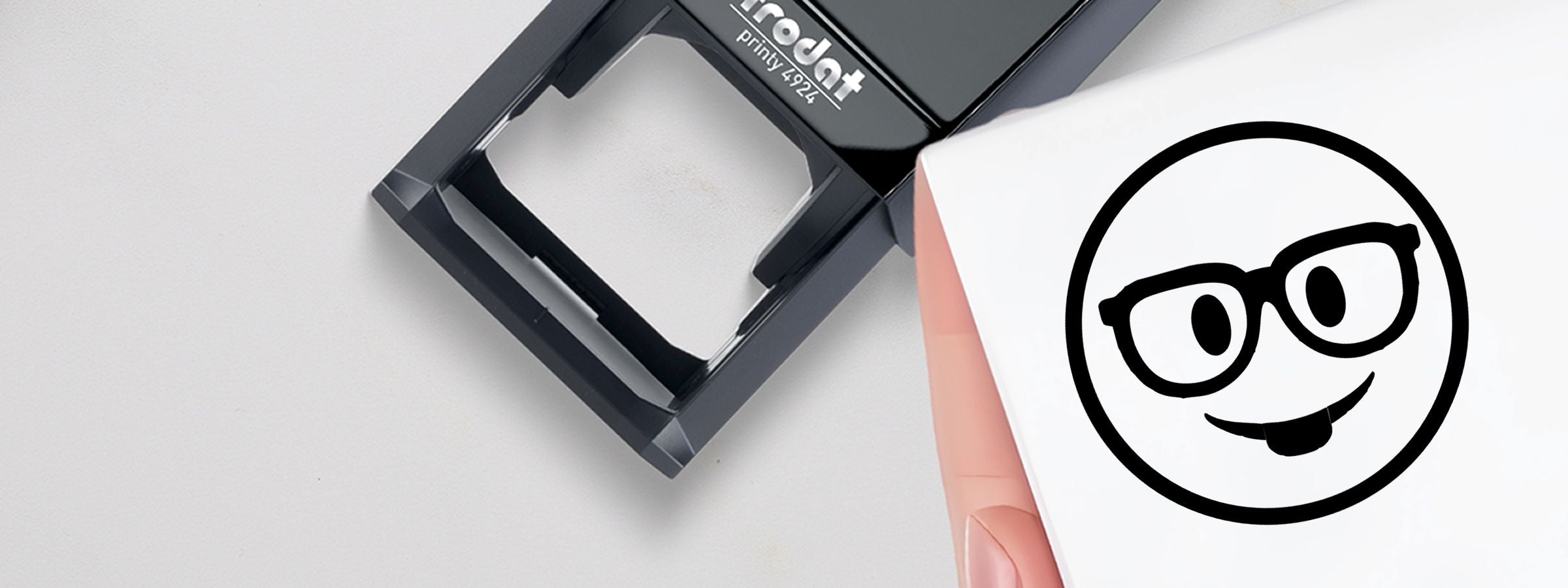 Black self-inking stamp beside a white sheet with a cartoon face wearing glasses; Stock Designer Stamps.