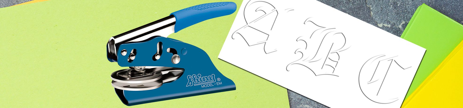 Blue handheld embossing tool with Monogram Embossers on colorful paper, showing embossed letter designs on white sheet.