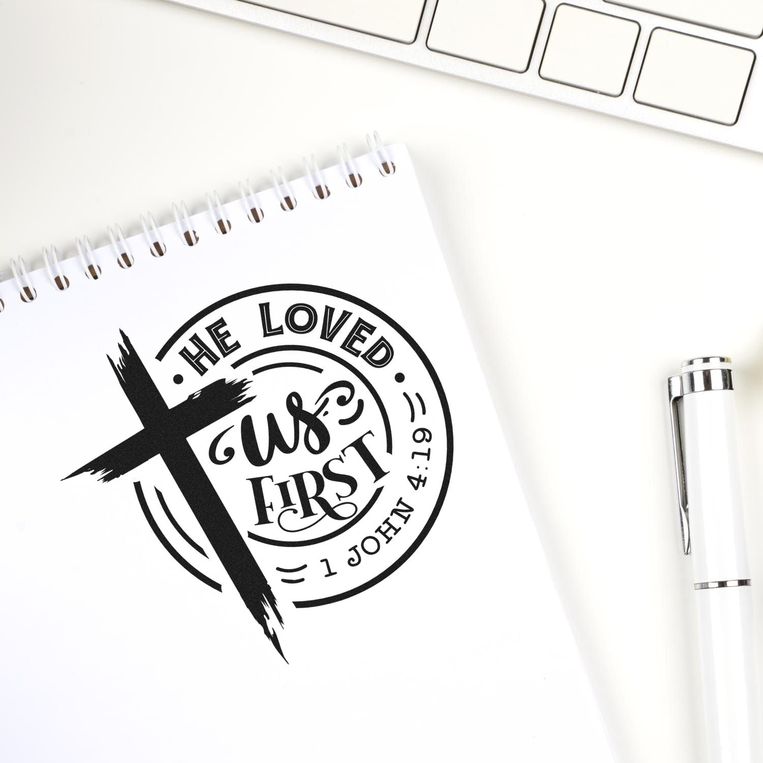 Christian rubber stamp design with cross and He Loved Us First Bible verse John 4:19 on notebook, with pen and keyboard