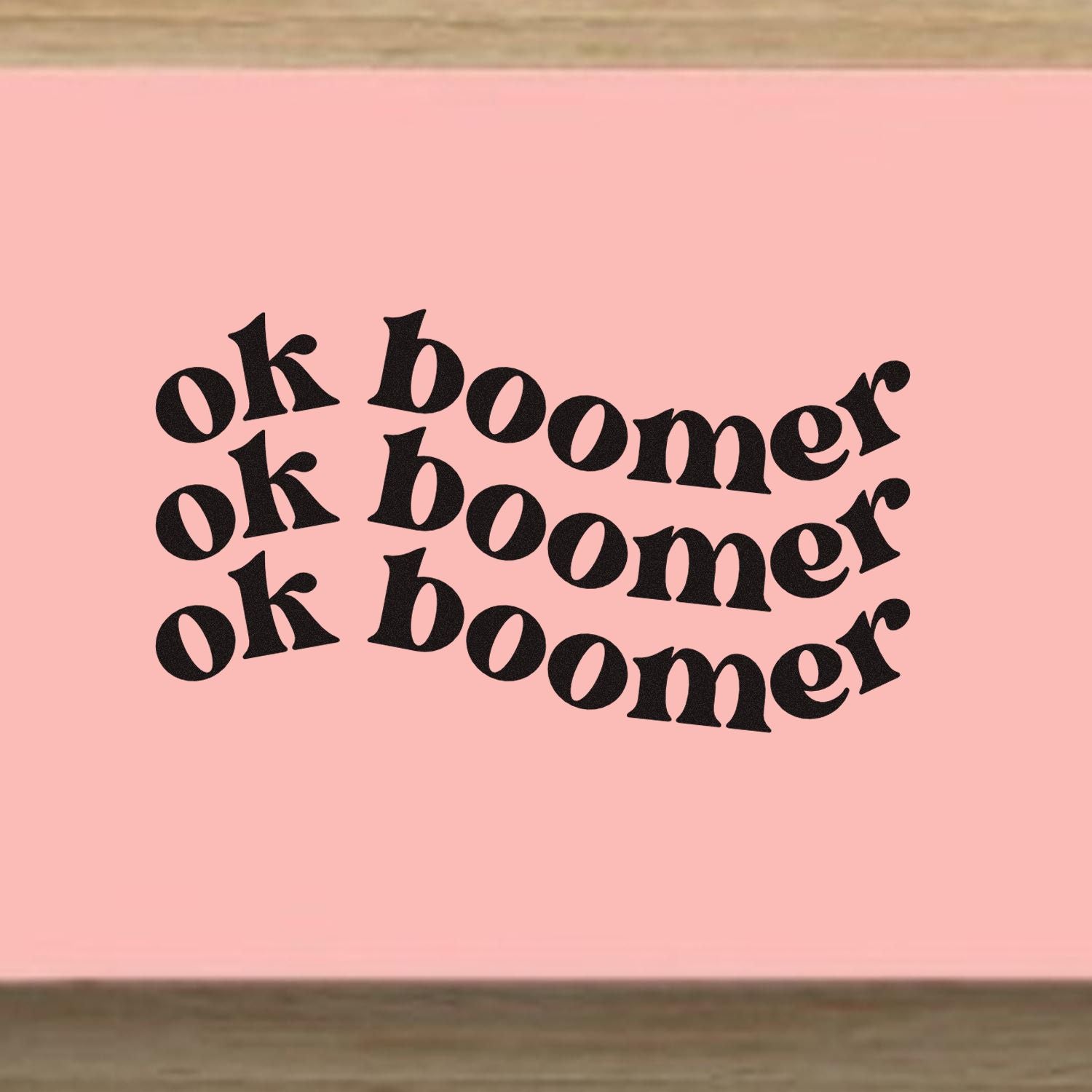 OK Boomer rubber stamp design with bold retro black text in three wavy lines on a pink background.