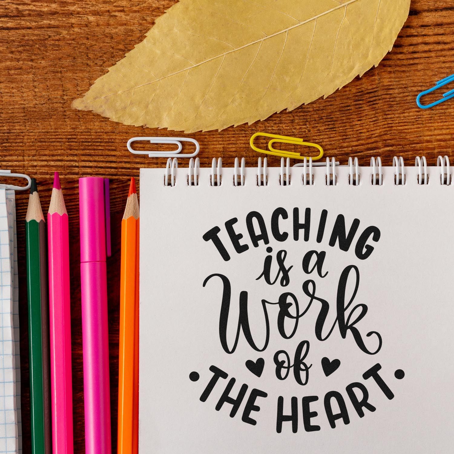 Teacher rubber stamp design with Teaching Is a Work of the Heart on a notebook, surrounded by pencils and paper clips.