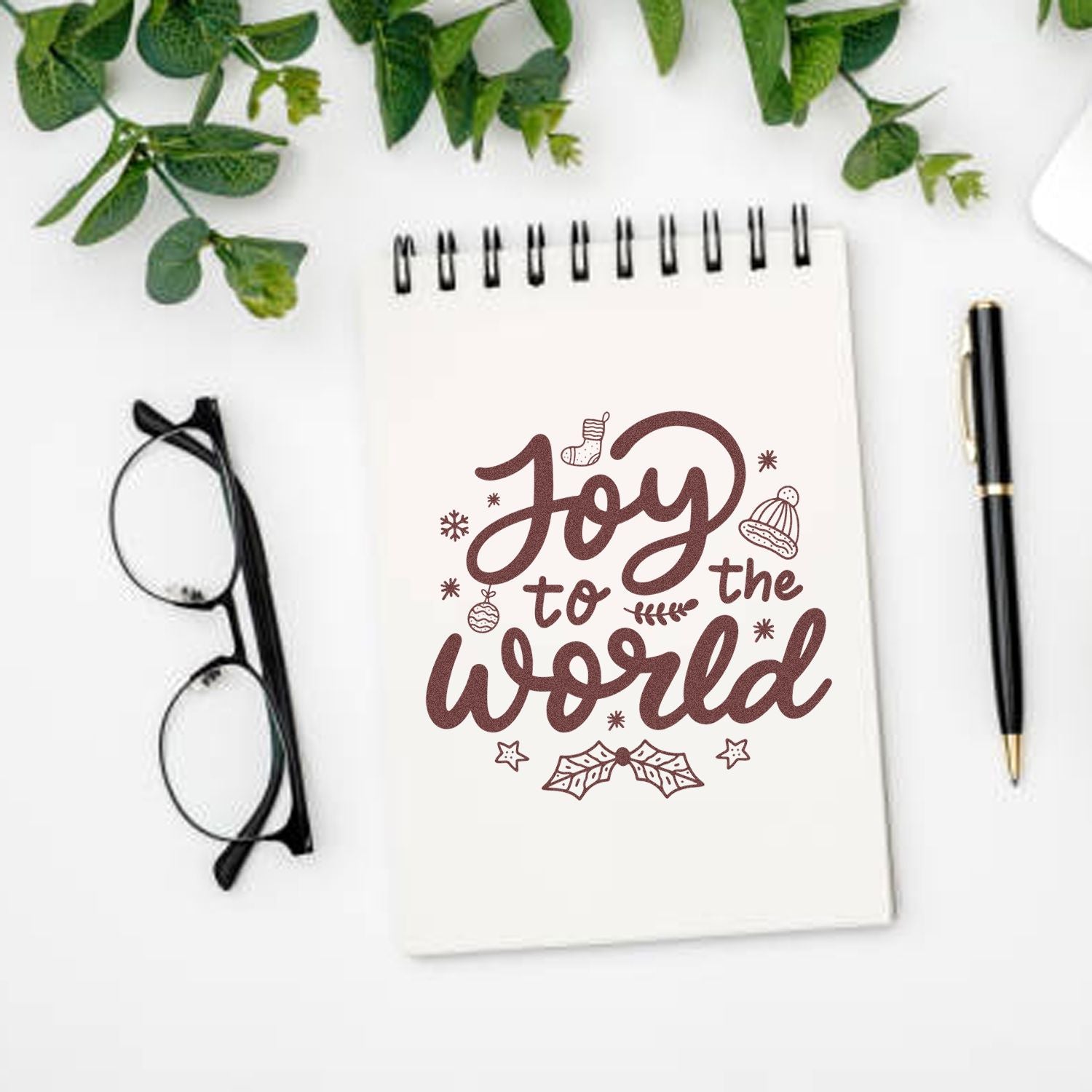 Joy to the World holiday rubber stamp design on notebook, with Christmas icons, pen, glasses, and greenery.