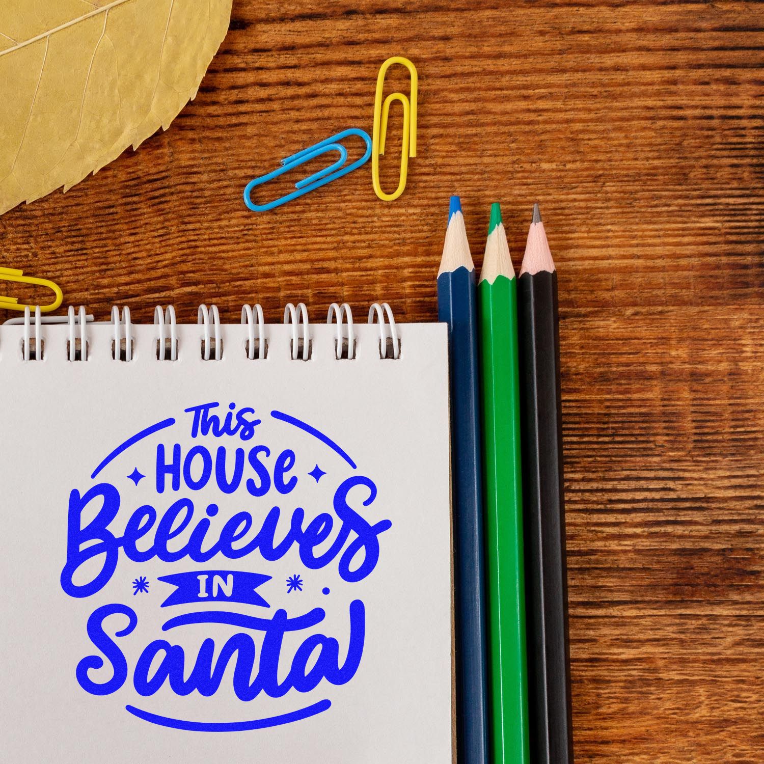 Christmas rubber stamp design reading This House Believes in Santa on a notebook with pencils and paper clips.
