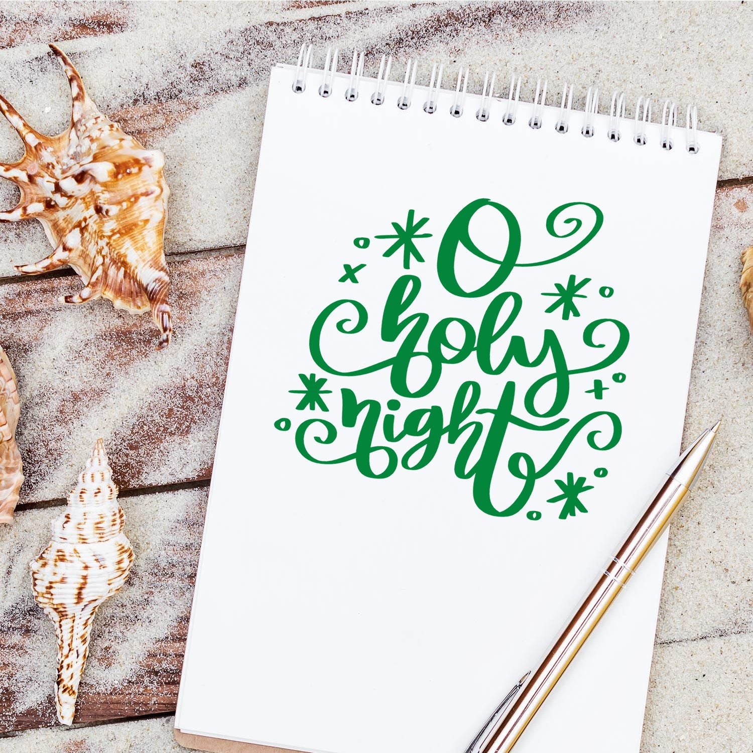 O Holy Night Christmas rubber stamp design in green script on a notebook, styled with seashells for coastal holiday crafts.