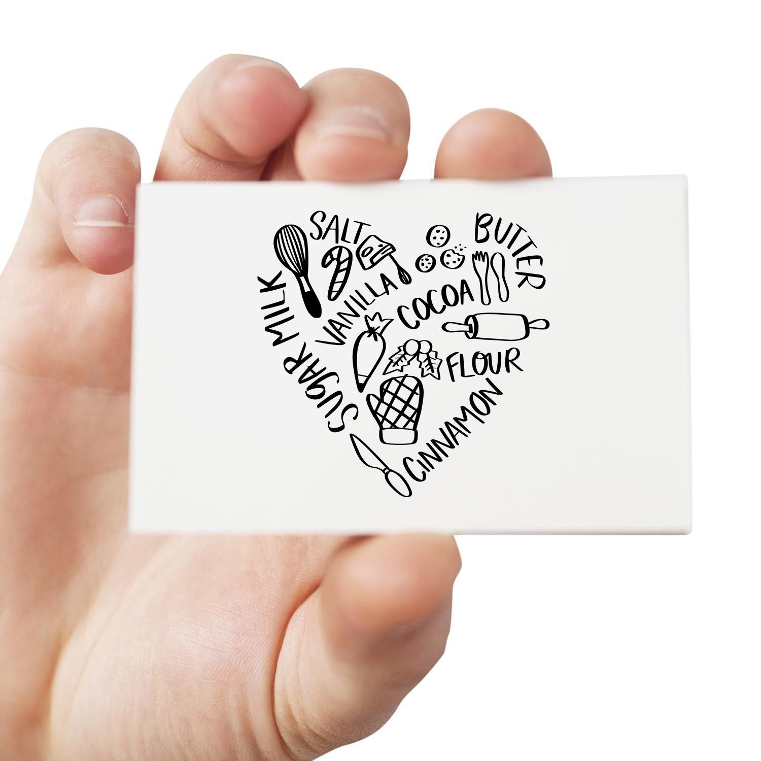 Hand holding baking ingredients heart rubber stamp featuring salt, butter, milk, sugar, vanilla, cocoa, flour and cinnamon.