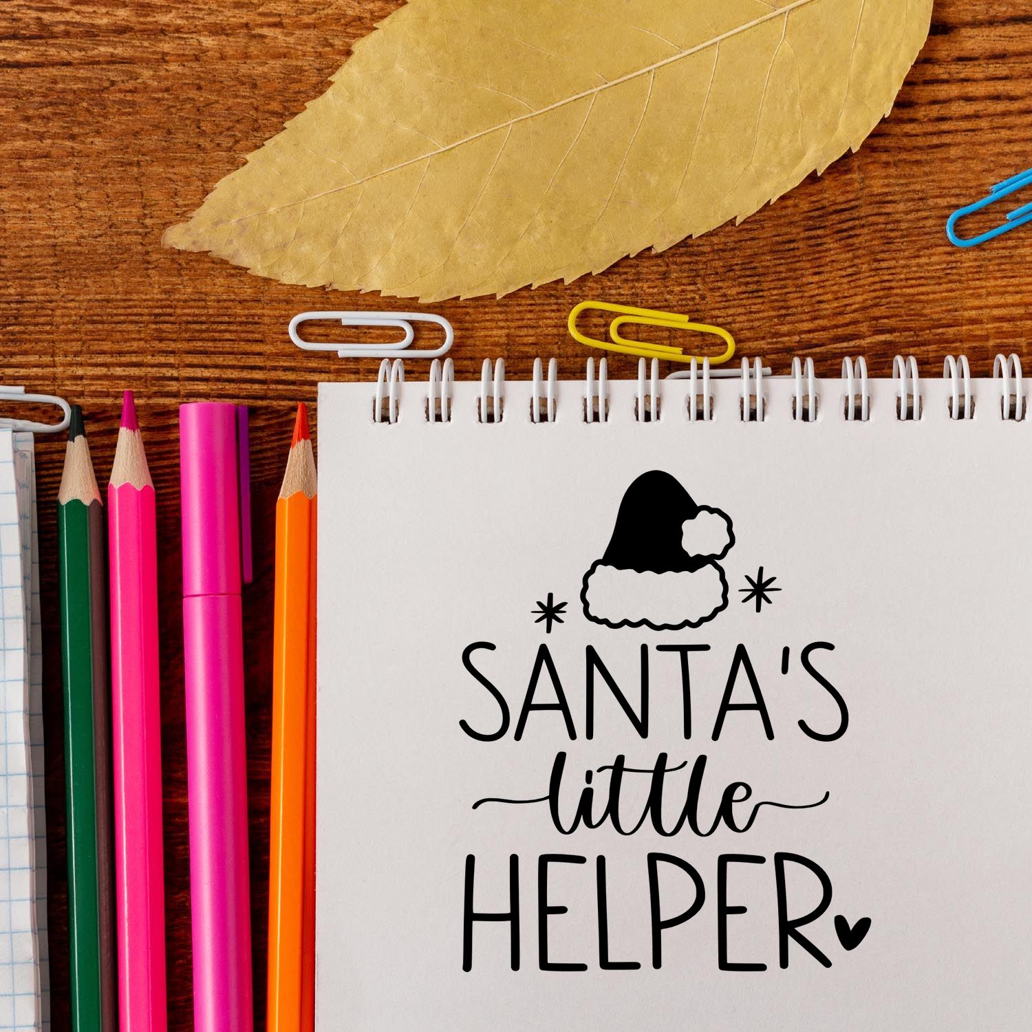 Holiday rubber stamp mockup with Santas little helper design on notebook, surrounded by pencils and paper clips.
