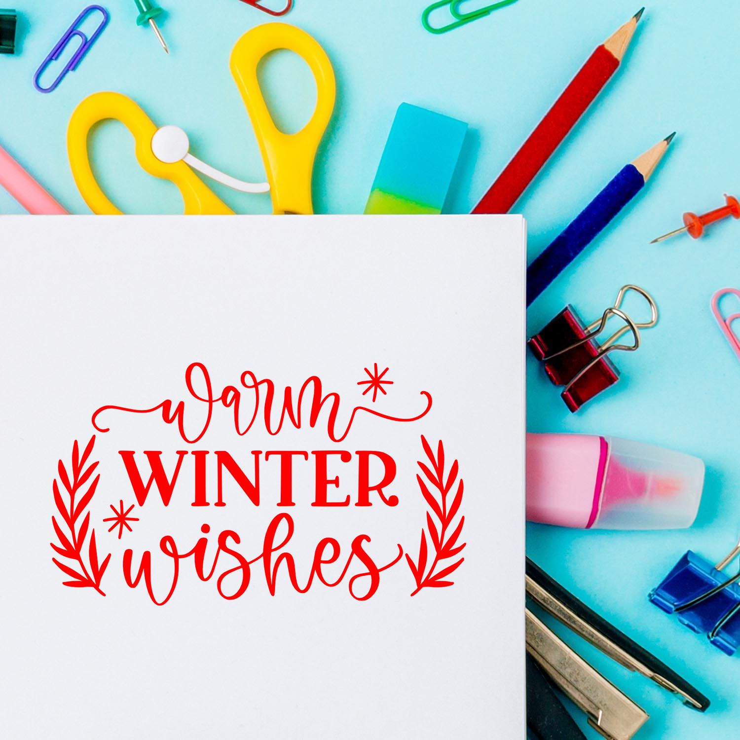 Warm winter wishes rubber stamp impression on white paper, surrounded by colorful stationery supplies.