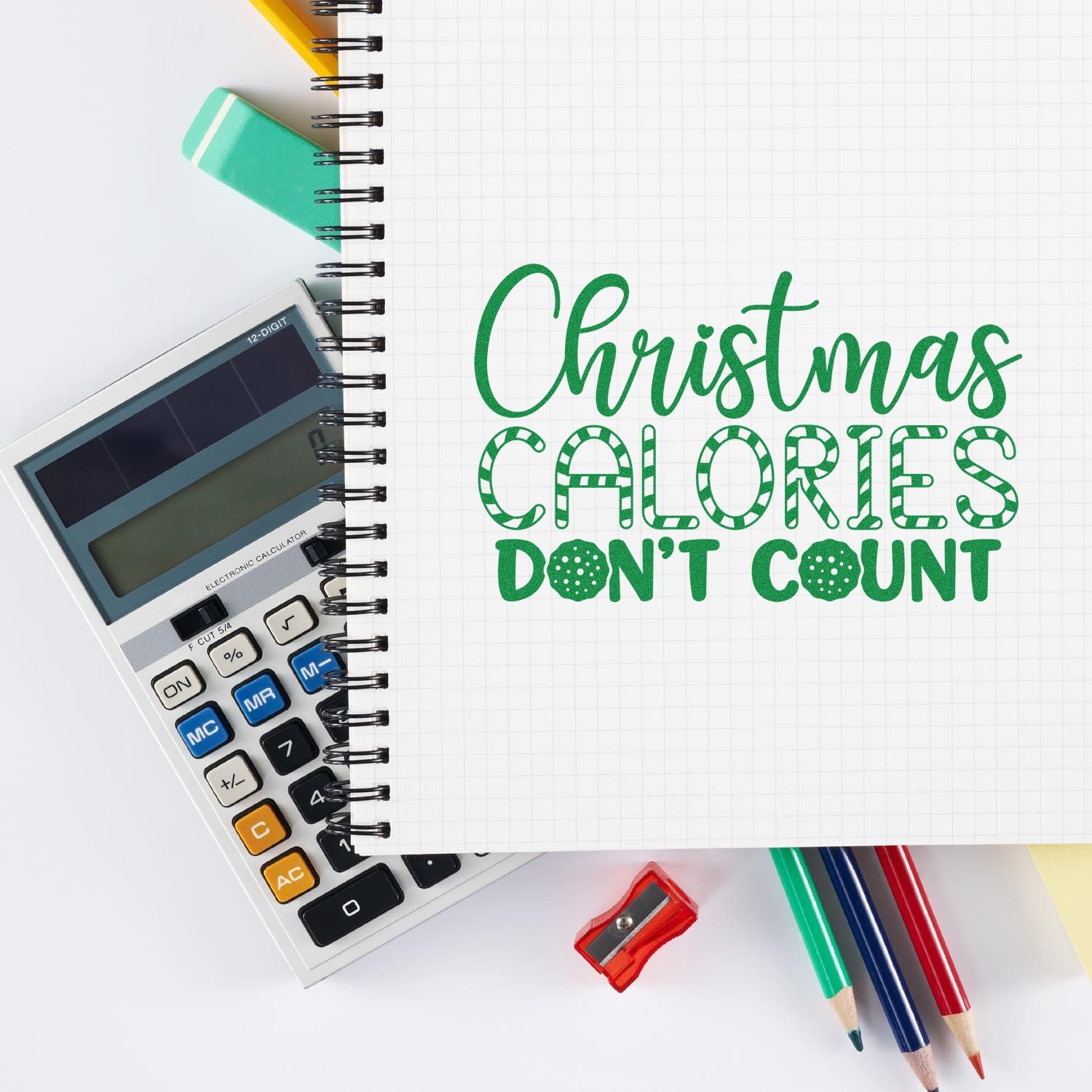 Christmas rubber stamp design with green Christmas calories don't count lettering on notebook beside calculator and pencils.
