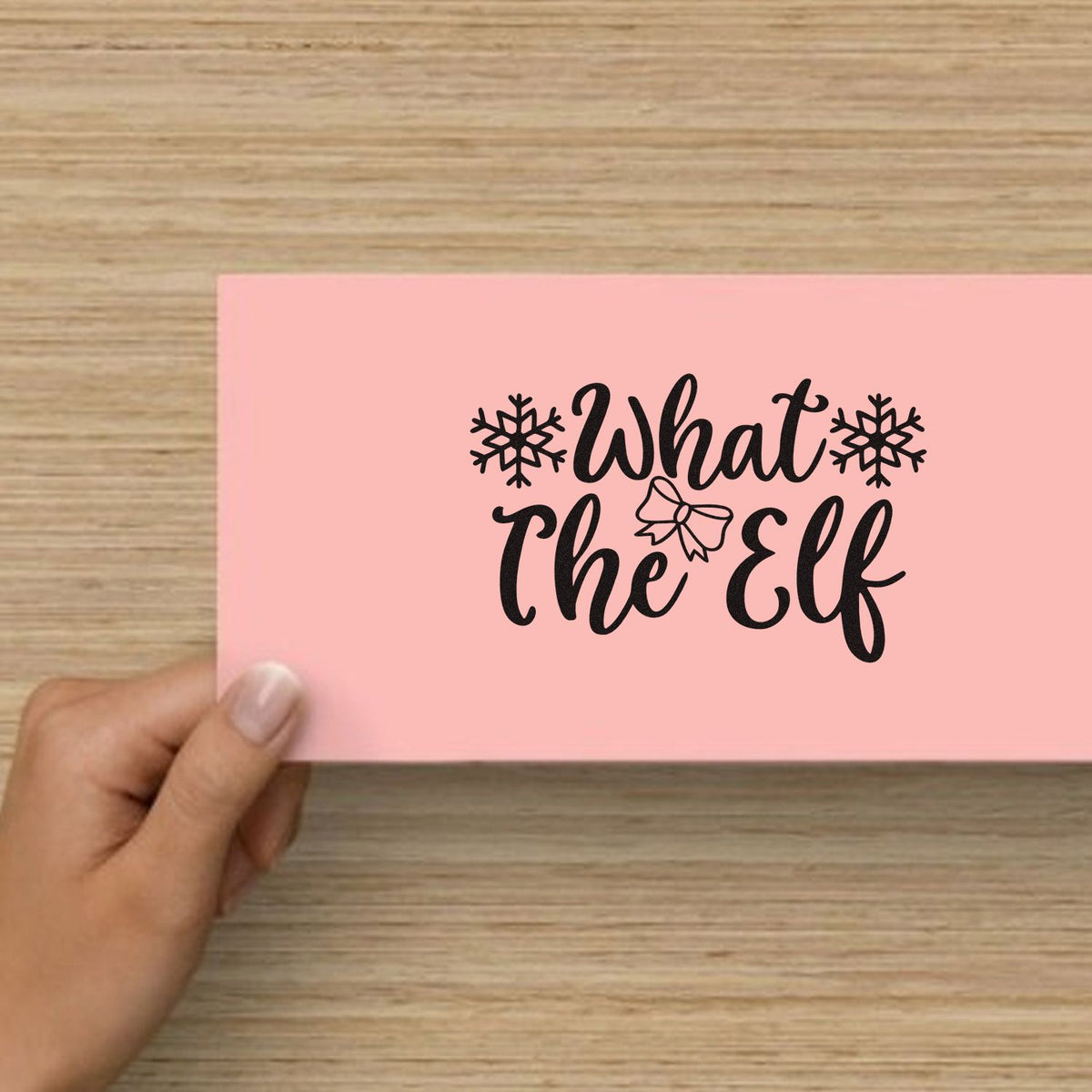 What The Elf Snowflakes Premium Wooden Stamp - Vintage Pro imprint stamped in black ink. "What the Elf" is framed by snowflakes with a small bow between the words.