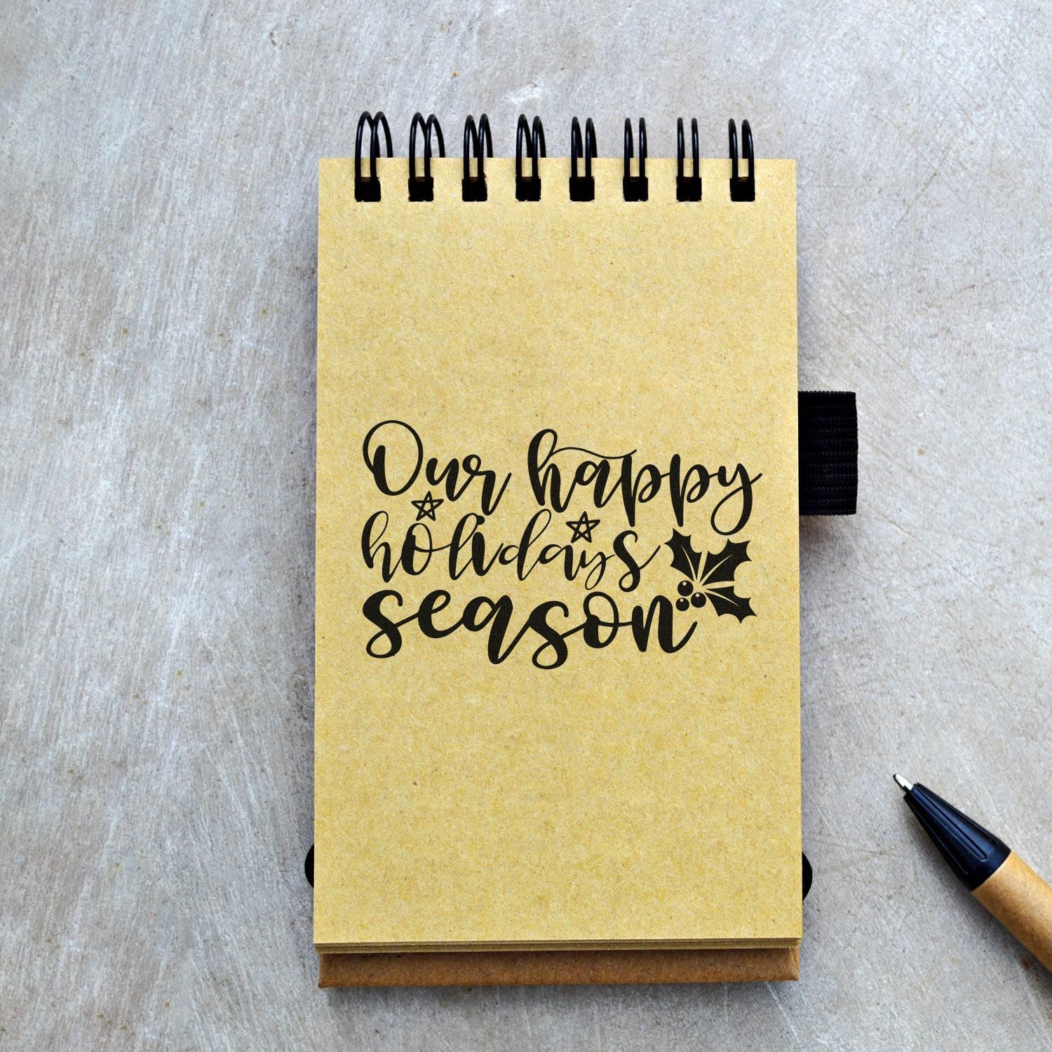 Holiday rubber stamp design reading Our Happy Holidays Season with holly on a kraft notebook mockup.