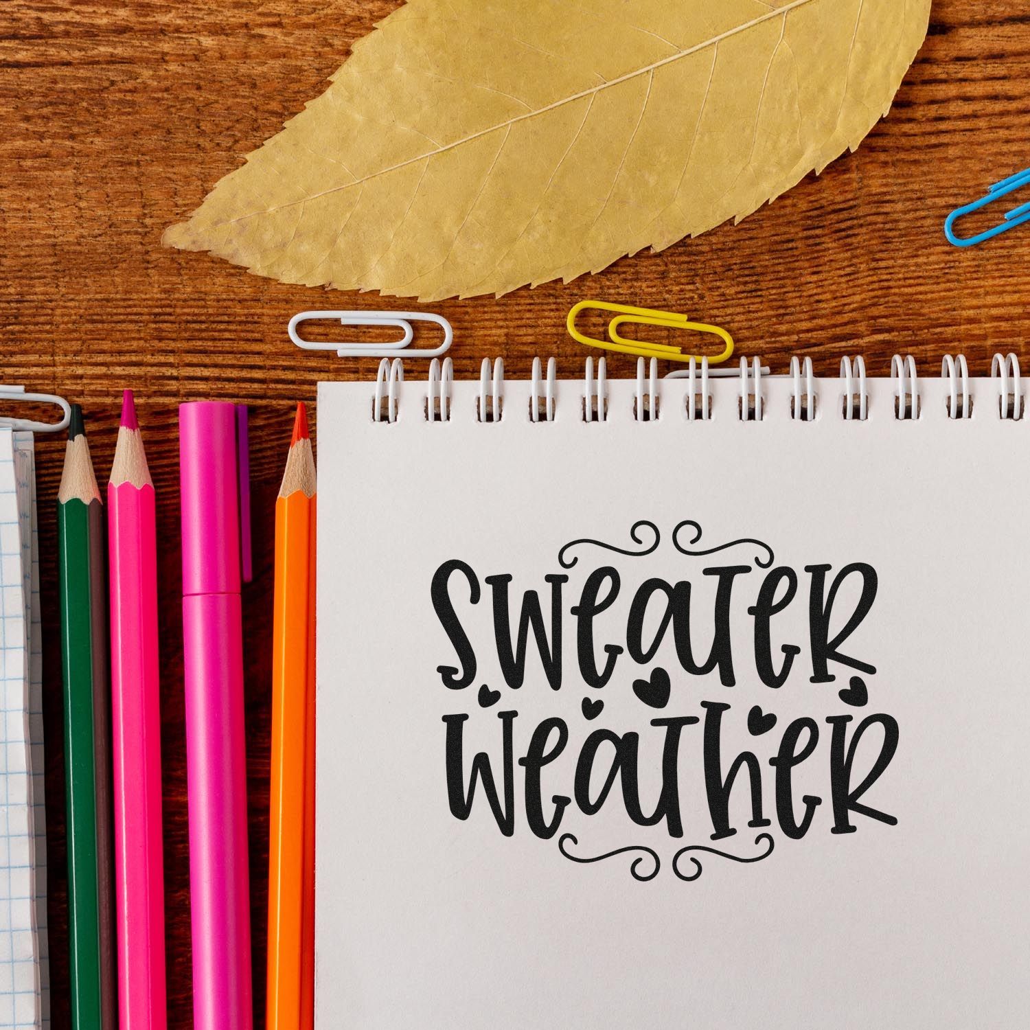 Sweater Weather rubber stamp design on a notebook, styled with pencils and a fall leaf for autumn journaling and crafts.