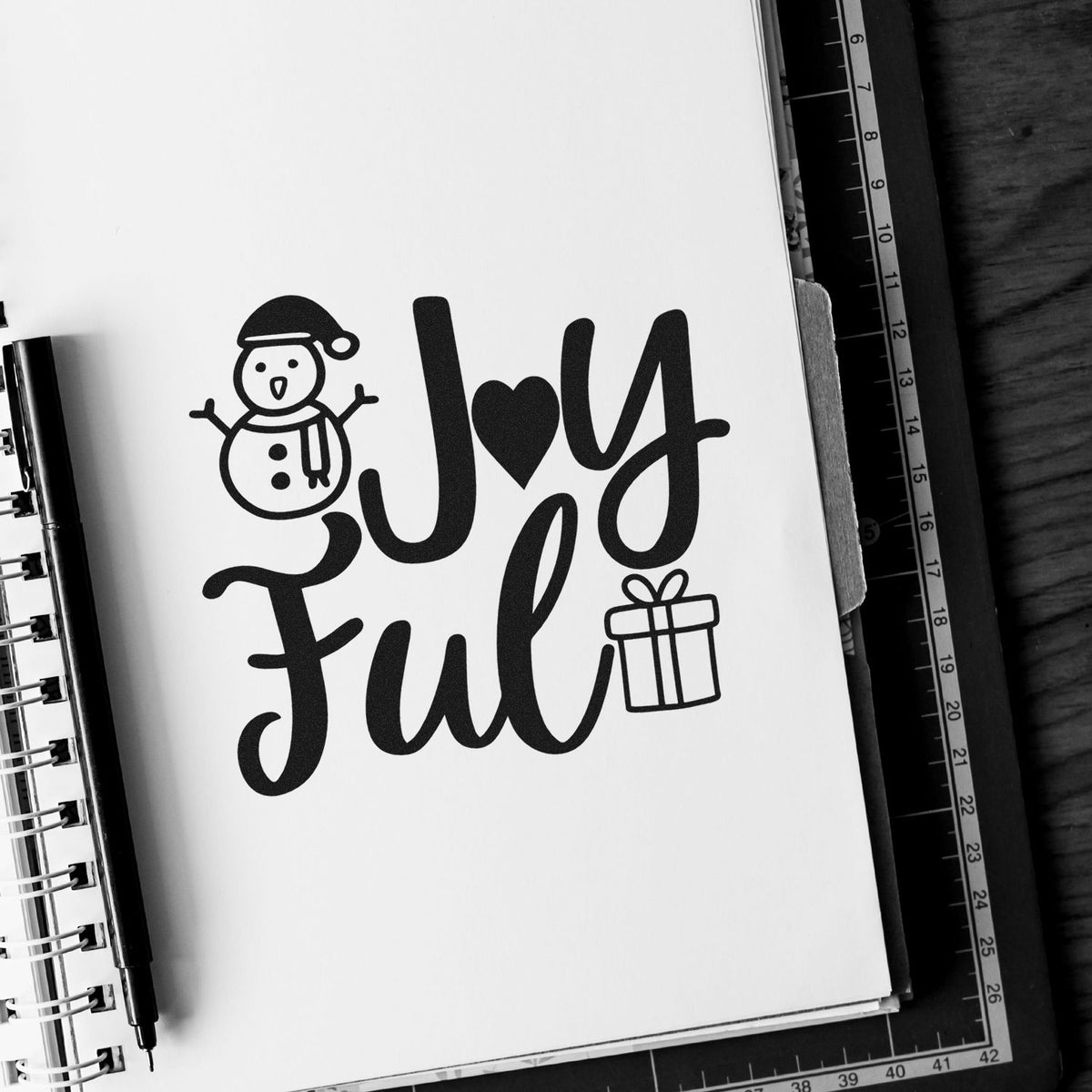 Joyful Snowman And Gift Premium Wooden Stamp - Vintage Pro imprint stamped in black ink. "Joy Ful" features a heart replacing the O, a smiling snowman, and a gift.