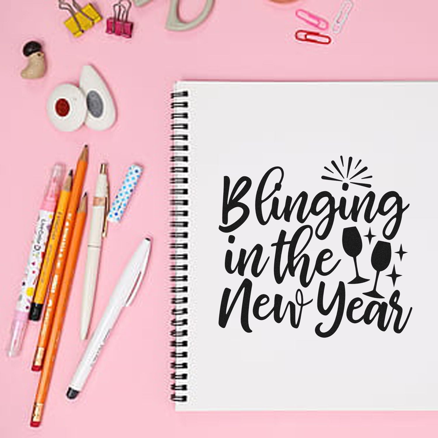 New Year rubber stamp design on spiral notebook with pens and office supplies on pink desk for festive stationery projects.