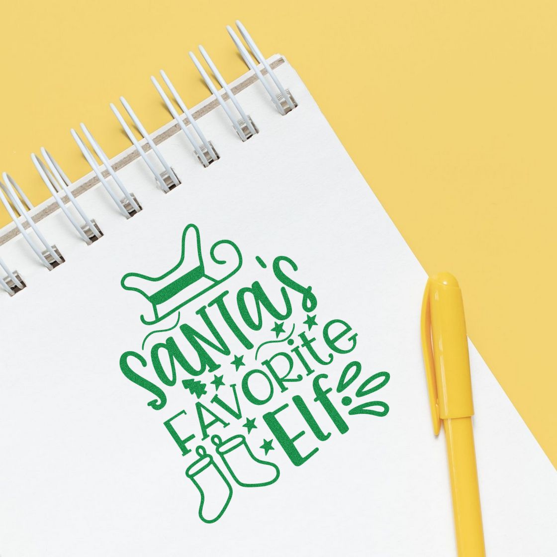Christmas rubber stamp design reading Santas Favorite Elf on a notebook with a yellow pen.