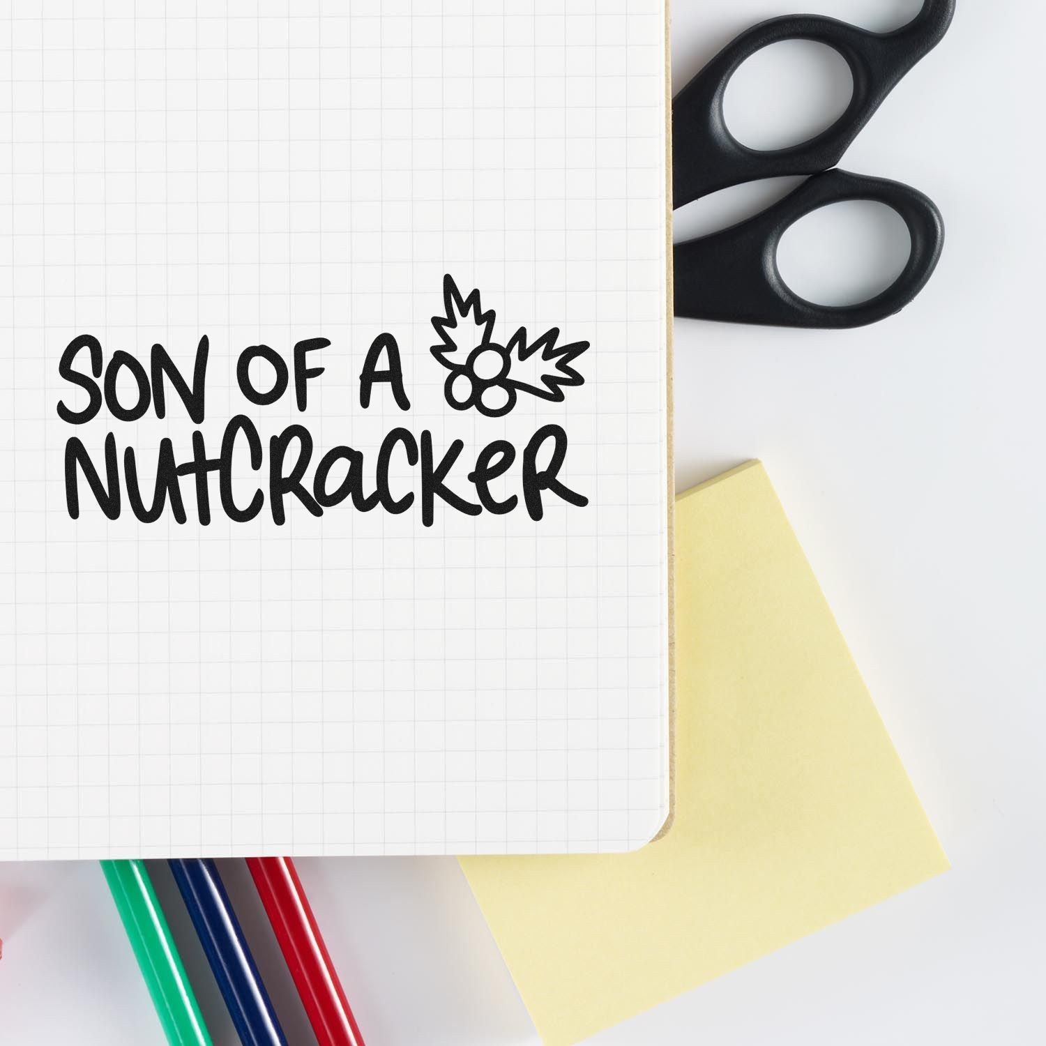 Son of a Nutcracker holiday rubber stamp design on notebook, with holly accent and craft supplies for Christmas projects.