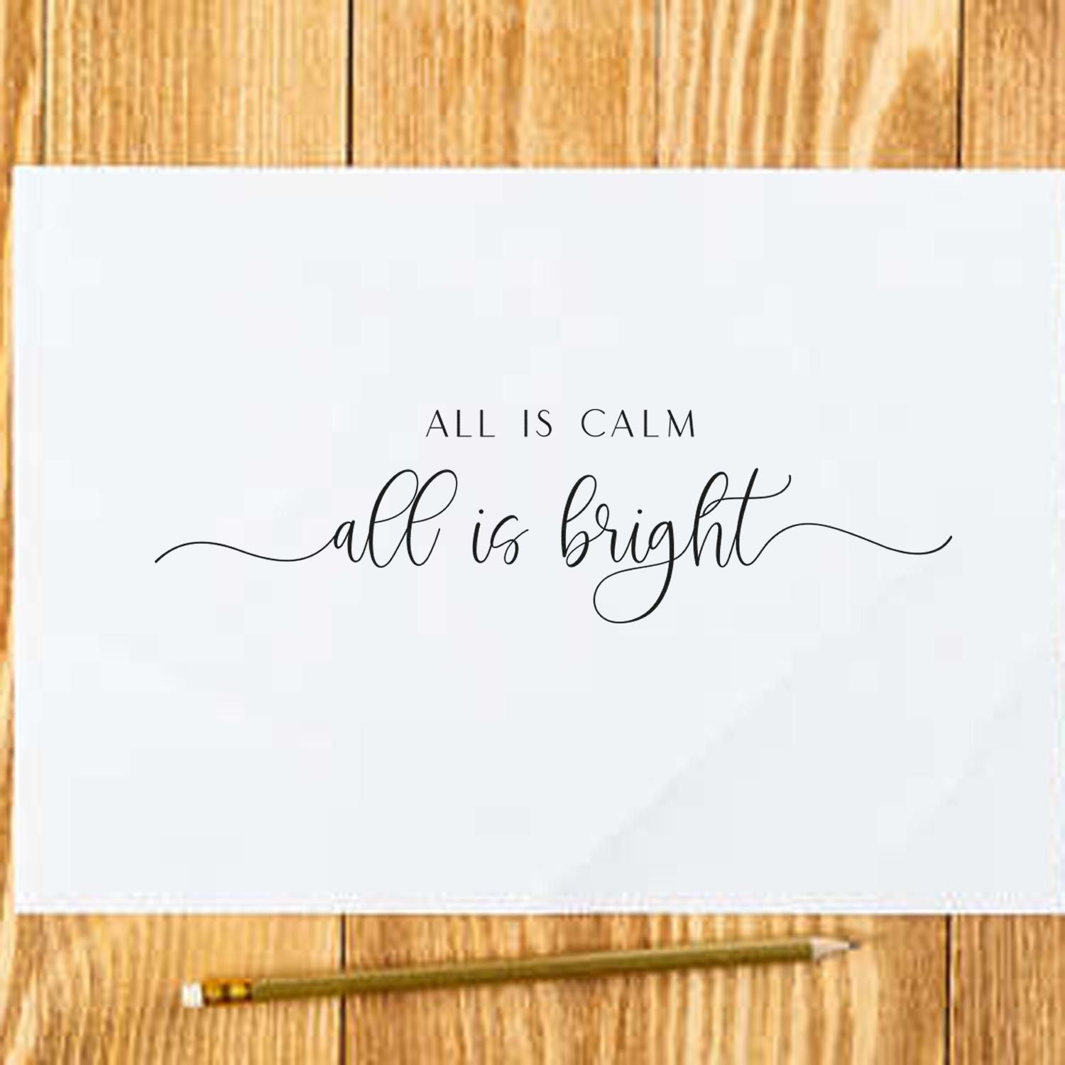 Holiday rubber stamp design reading All is calm, all is bright on white paper with pencil on wood background.