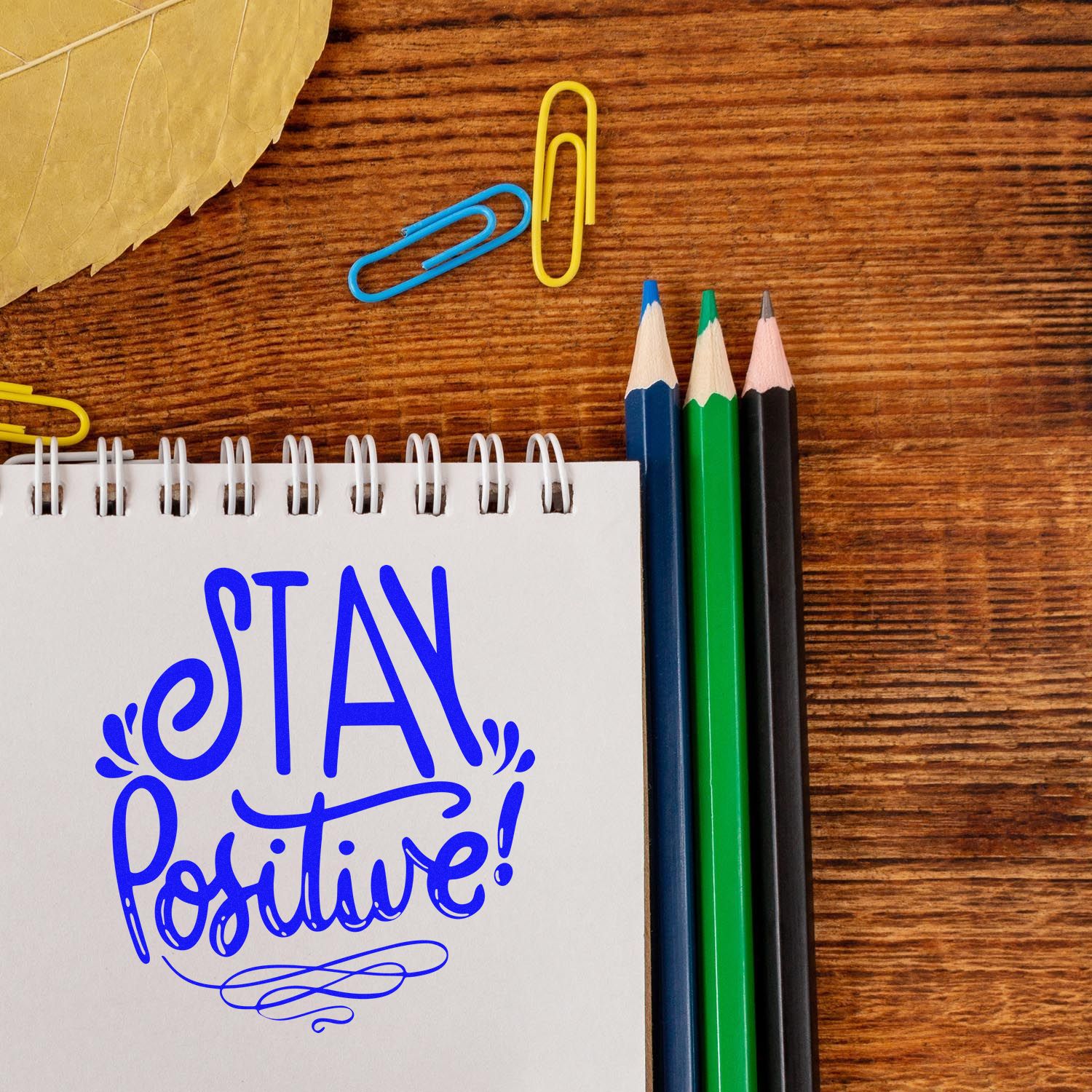 Motivational Stay Positive rubber stamp design on notebook, with pencils and paper clips on a wooden desk.