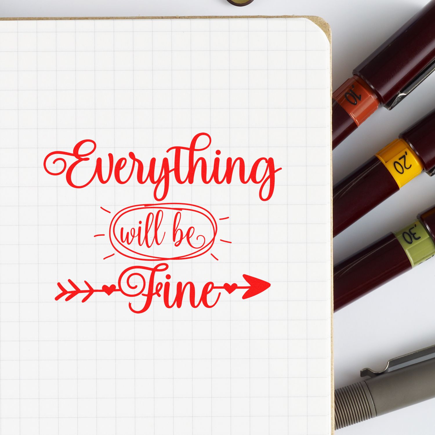 Motivational rubber stamp reading Everything will be Fine stamped on notebook paper, with ink stamp pens beside it.