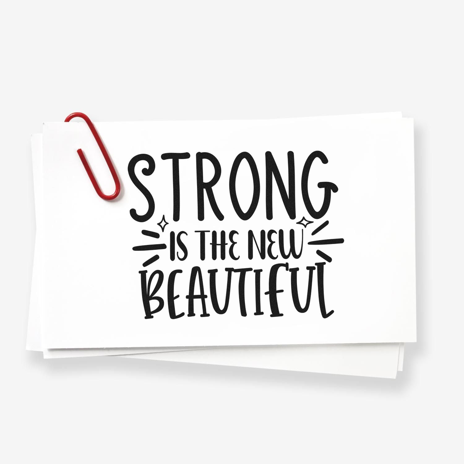 Strong Is the New Beautiful motivational rubber stamp design on stacked paper with red paperclip.