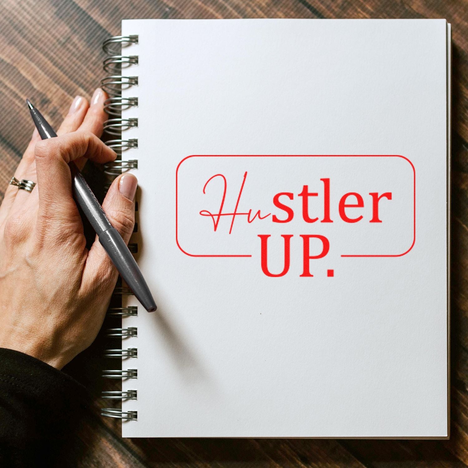 Custom rubber stamp reading Hustler Up on a spiral notebook, held by a hand with a pen for motivational branding.