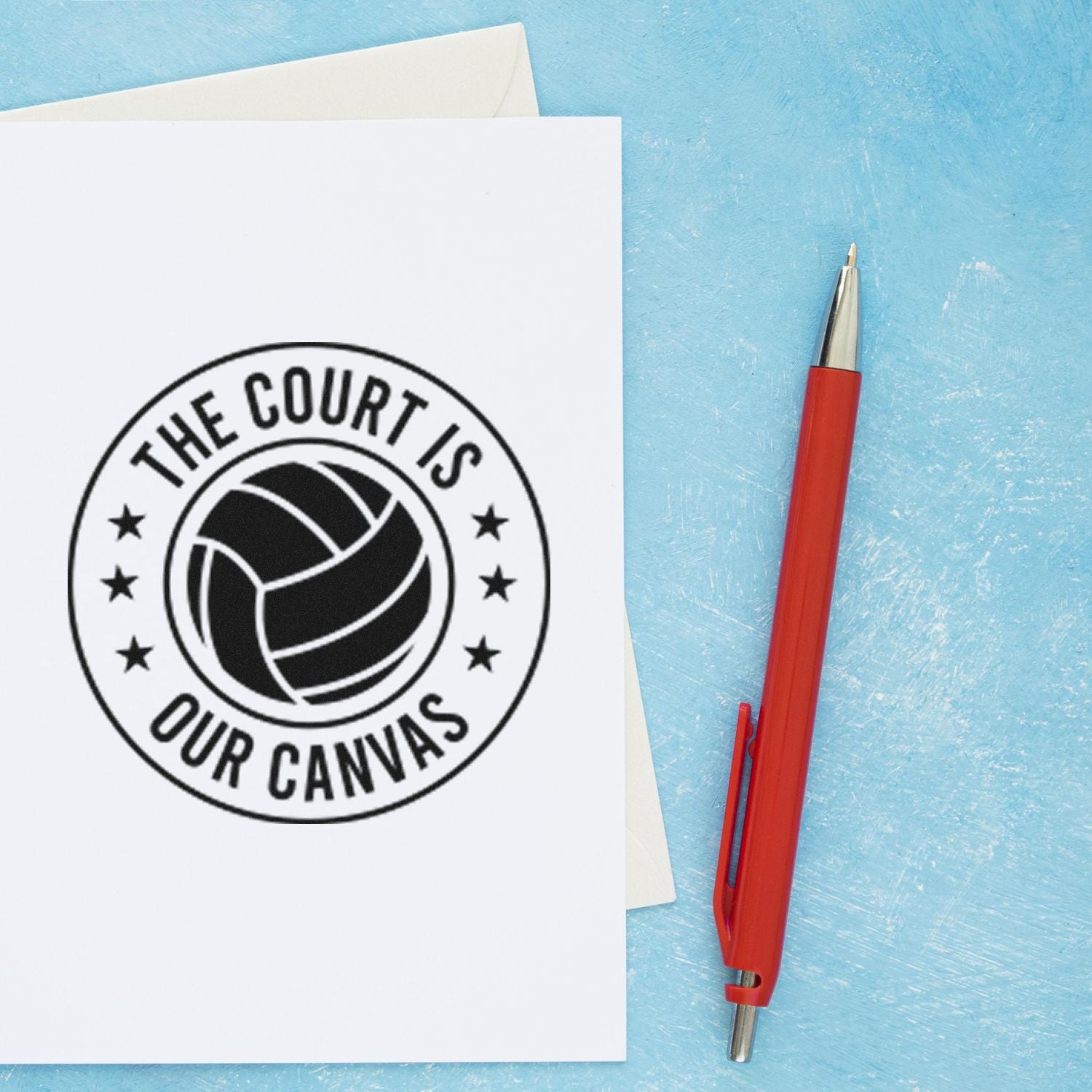 Custom rubber stamp with volleyball design and The Court Is Our Canvas imprint, shown beside a red pen.