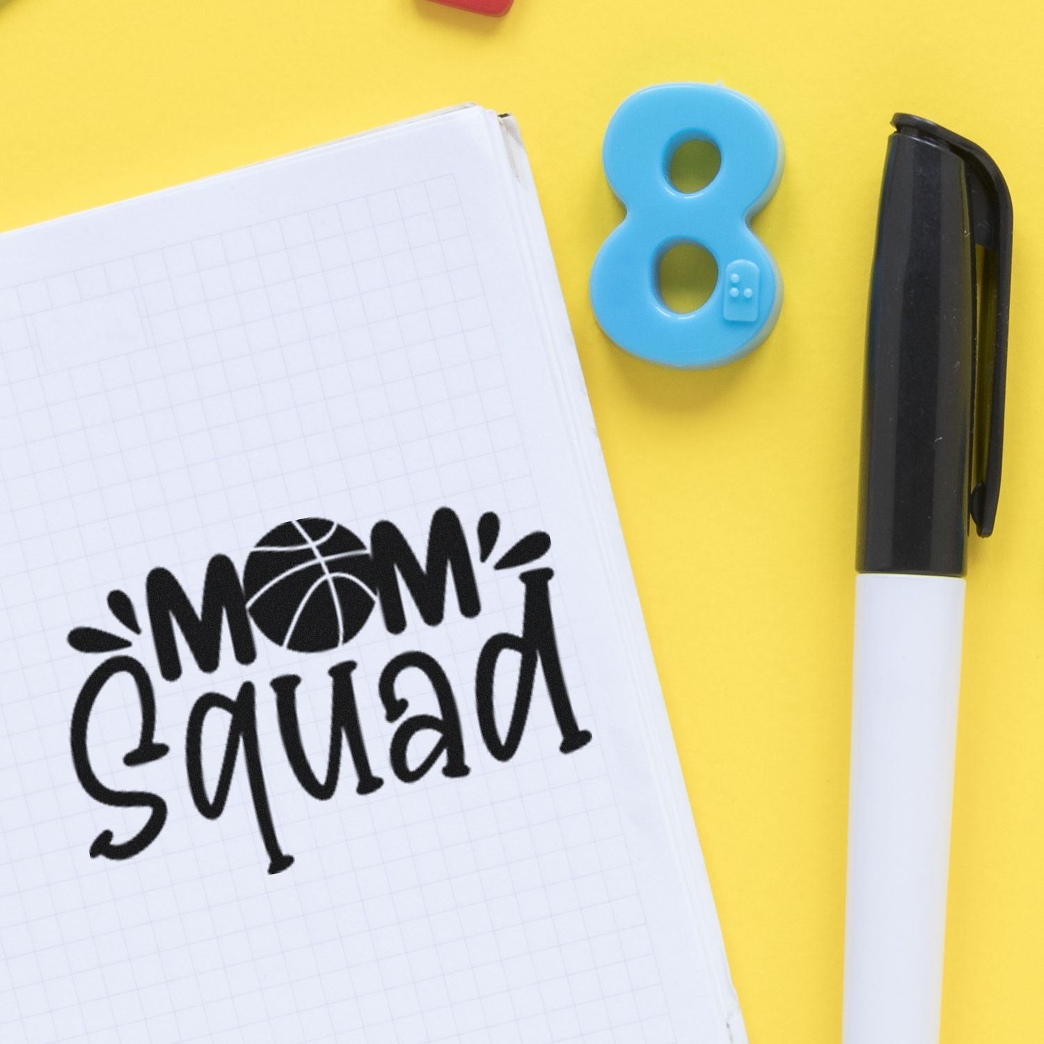 Custom rubber stamp imprint reading Mom Squad on notebook, with marker pen and school supplies on a yellow background.