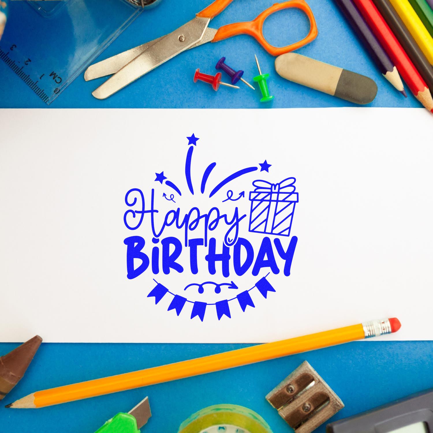 Happy Birthday rubber stamp imprint on white paper, framed by stationery and craft supplies on a blue desk.