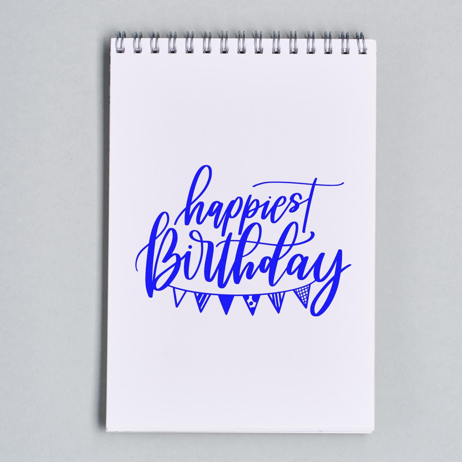 Blue happiest birthday rubber stamp design on a spiral notebook page with decorative bunting.